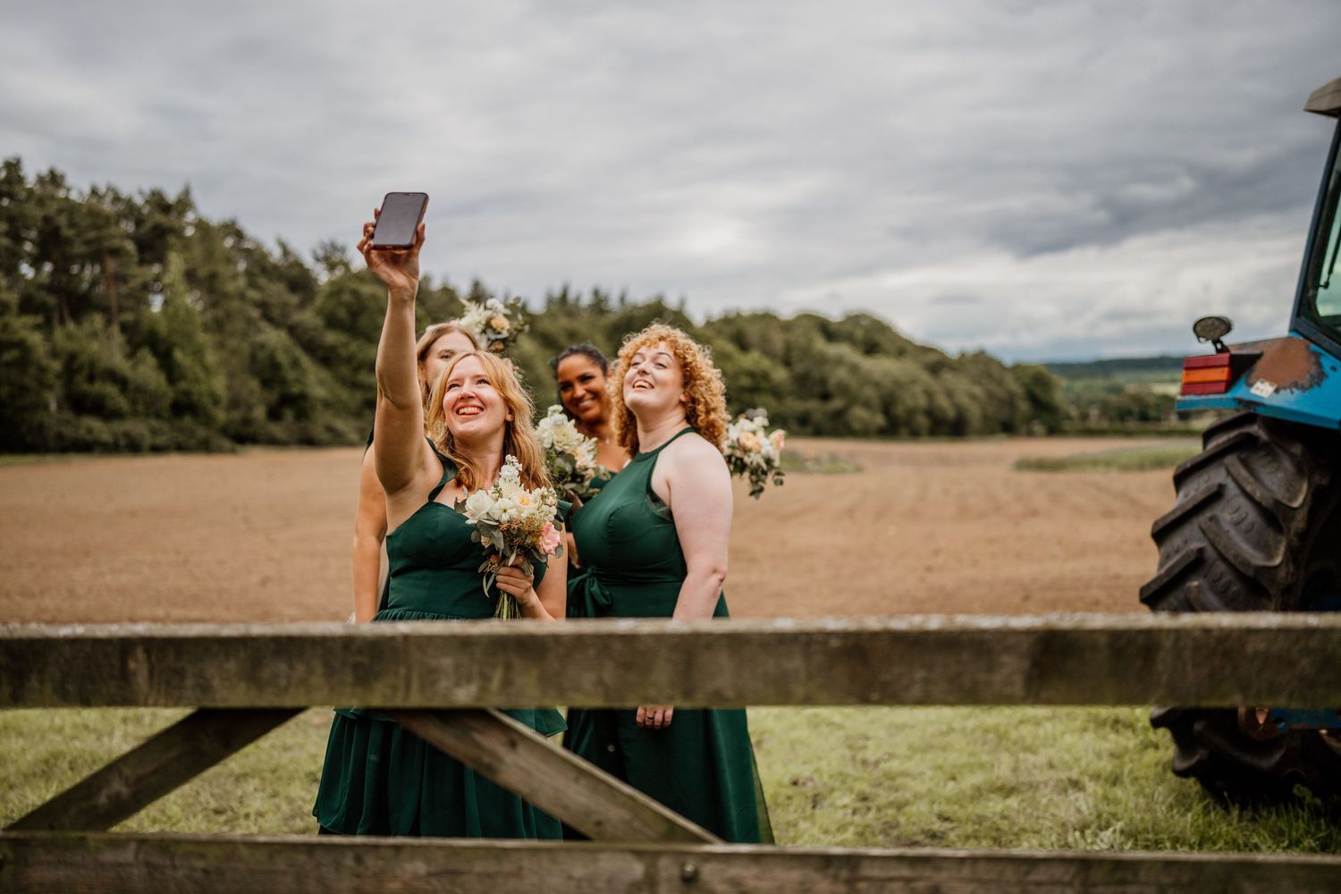Two bridesmaids in emerald green dresses laugh and celebrate together by a wooden fence in a countryside setting.