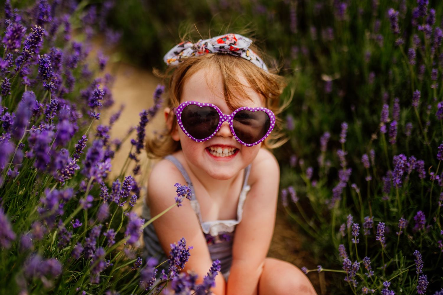 Smiling girl wearing heart-shaped sunglasses and floral headband sitting among purple lavender flowers in a field.