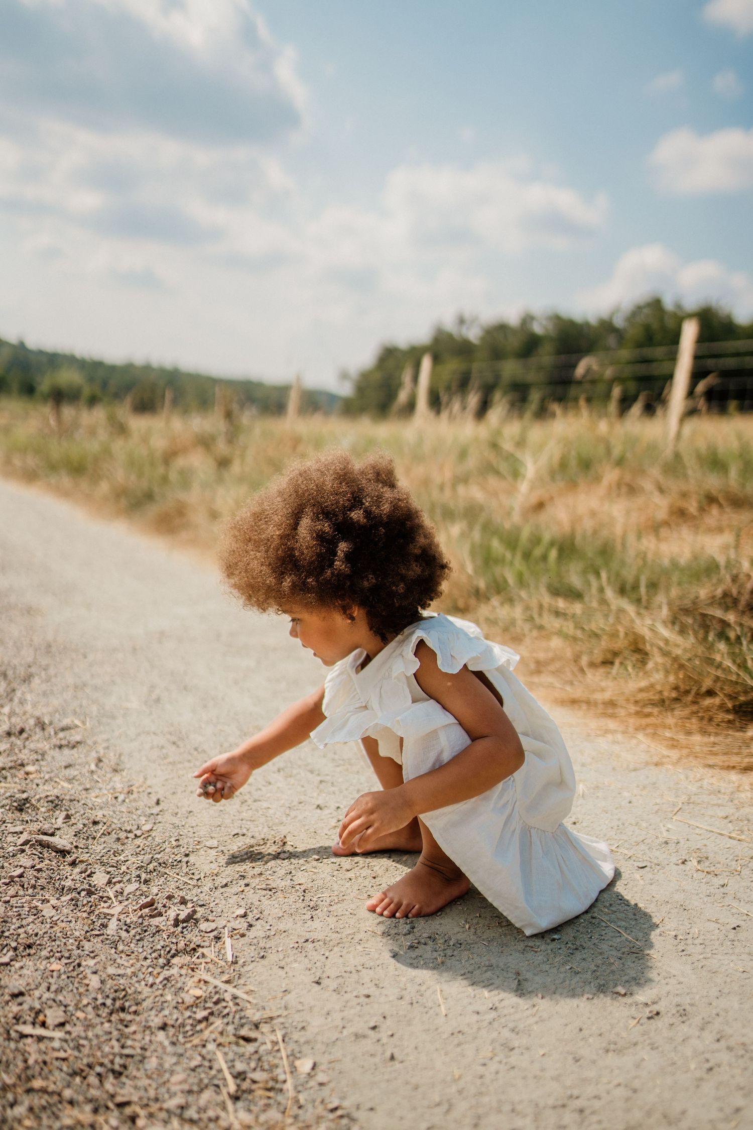 Child in white dress exploring dirt path on sunny summer day.