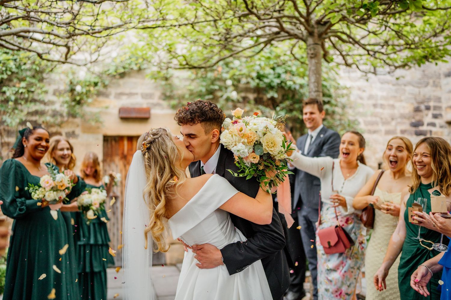 Wedding guests celebrate joyfully during outdoor garden reception with natural greenery and stone wall backdrop.