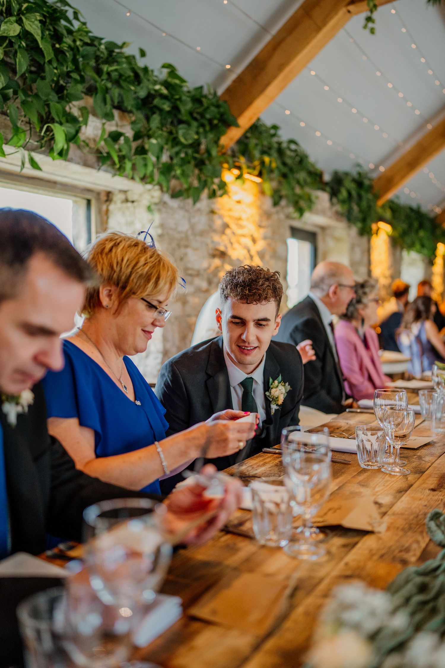 Wedding guests enjoy dinner at a long wooden table decorated with greenery and string lights.
