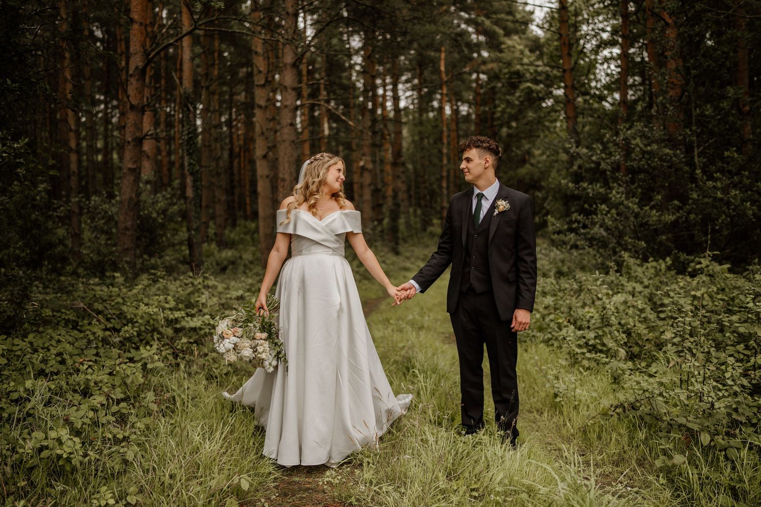 A couple in wedding attire holds hands while walking through a lush forest setting during golden hour.