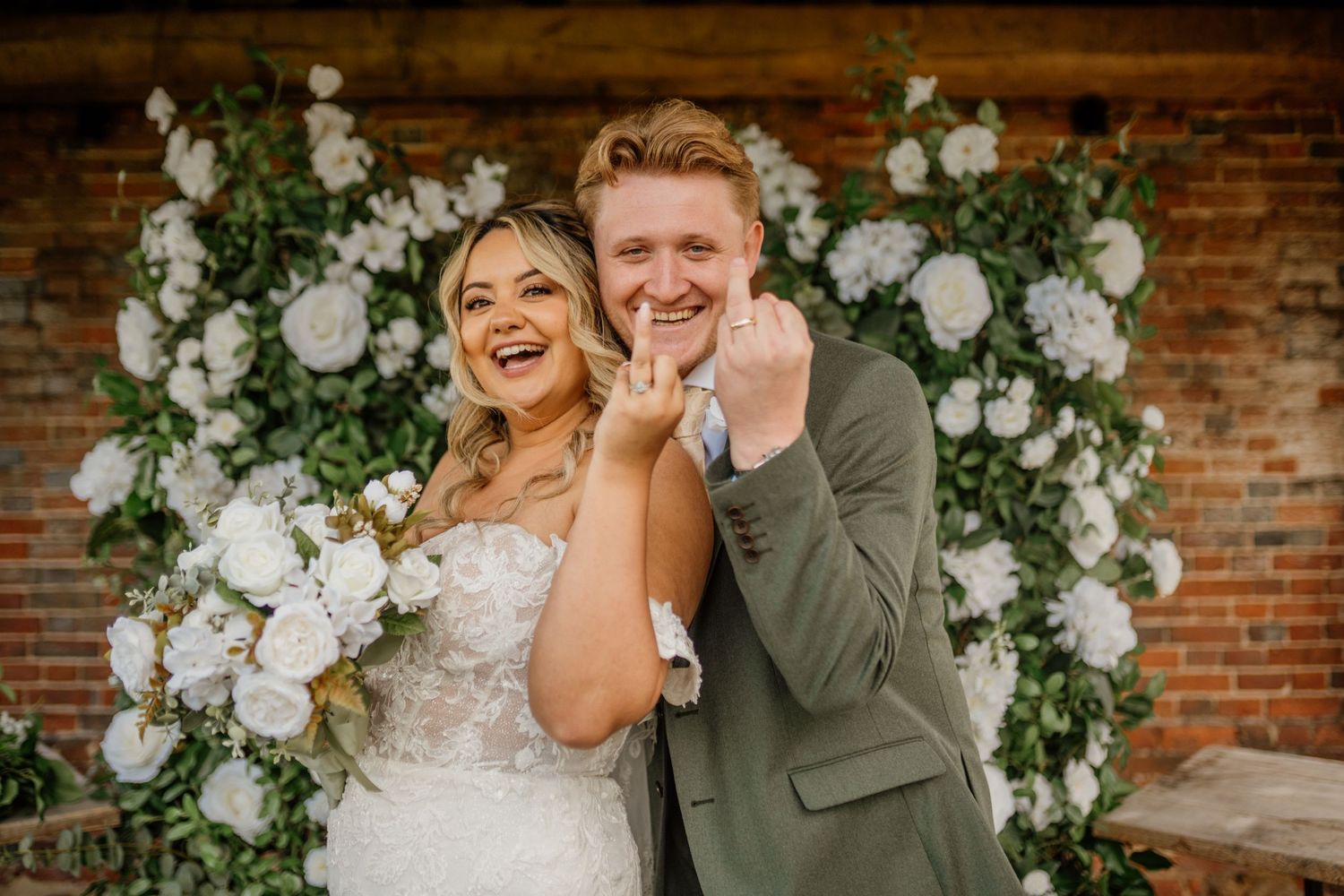 A joyful wedding couple laughs together in front of an elegant white floral arch against a brick wall.
