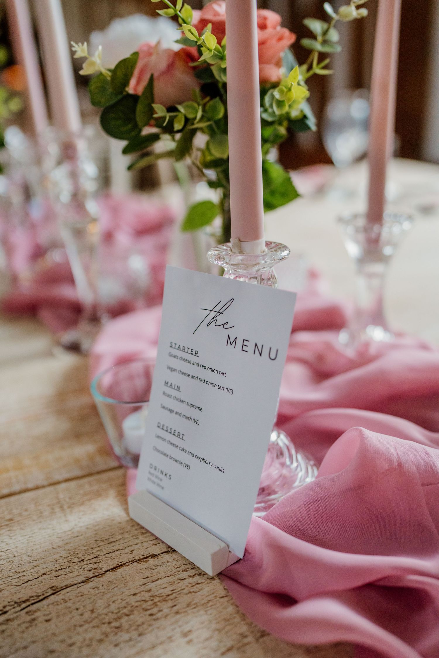 Wedding menu card displayed in crystal holder with pink taper candles and floral arrangements on table.
