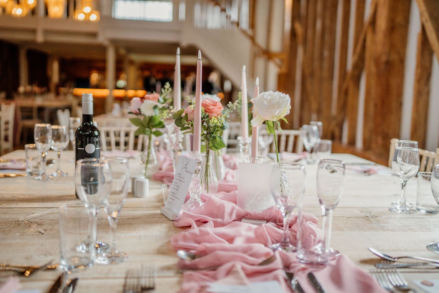 Elegant wedding reception table settings with pink napkins and white linens in a rustic barn venue.