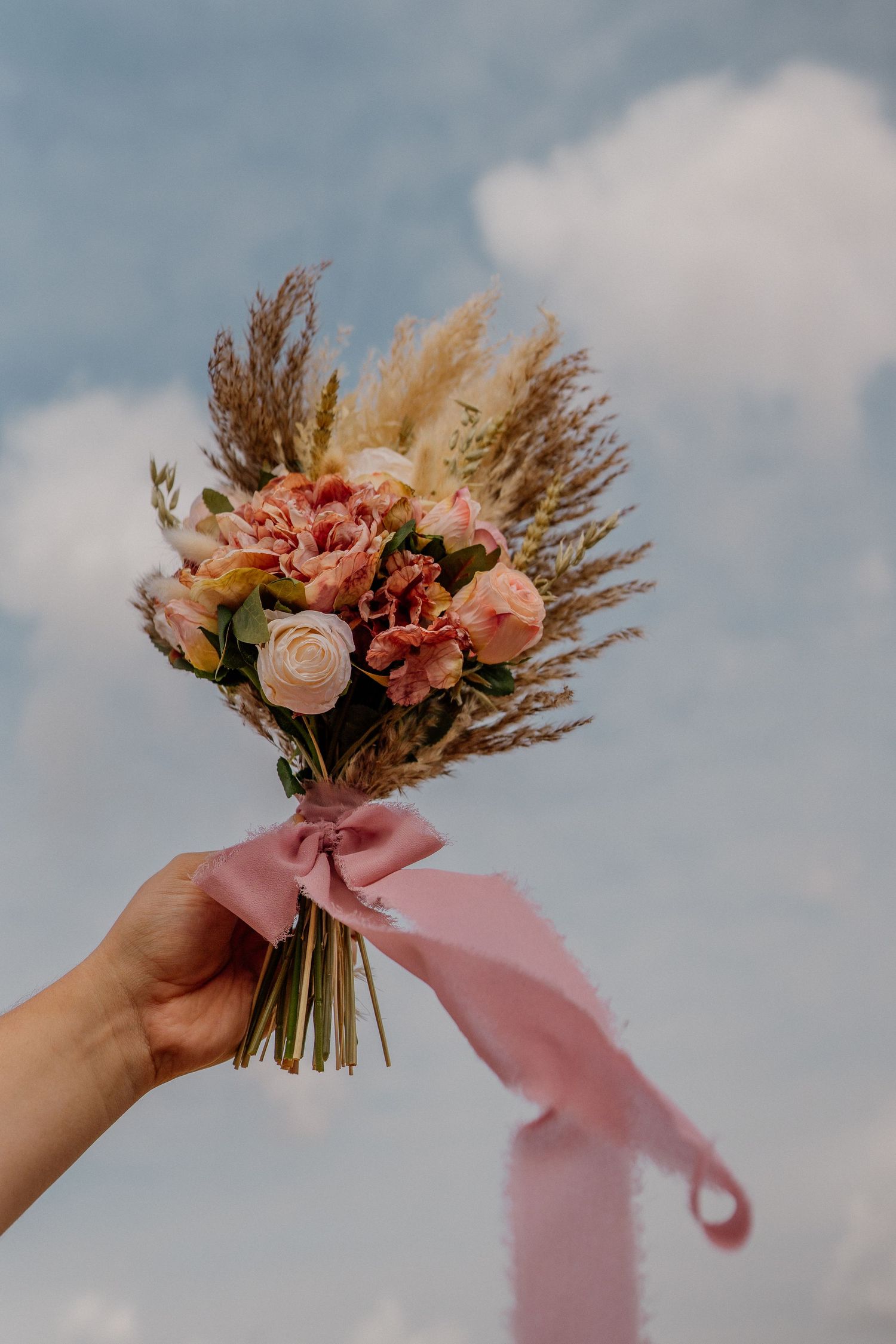 A delicate pink and peach floral bouquet with dried flowers held against a cloudy sky background.