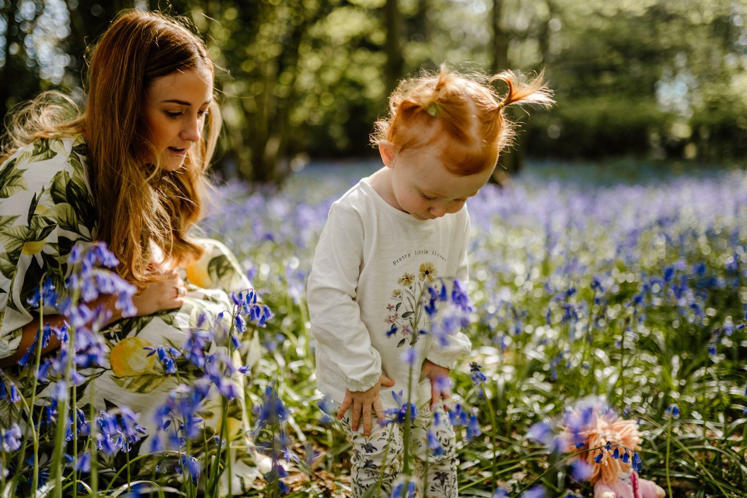 Two people sitting in a blooming field of purple bluebells in Hertfordshire surrounded by trees in springtime sunlight.