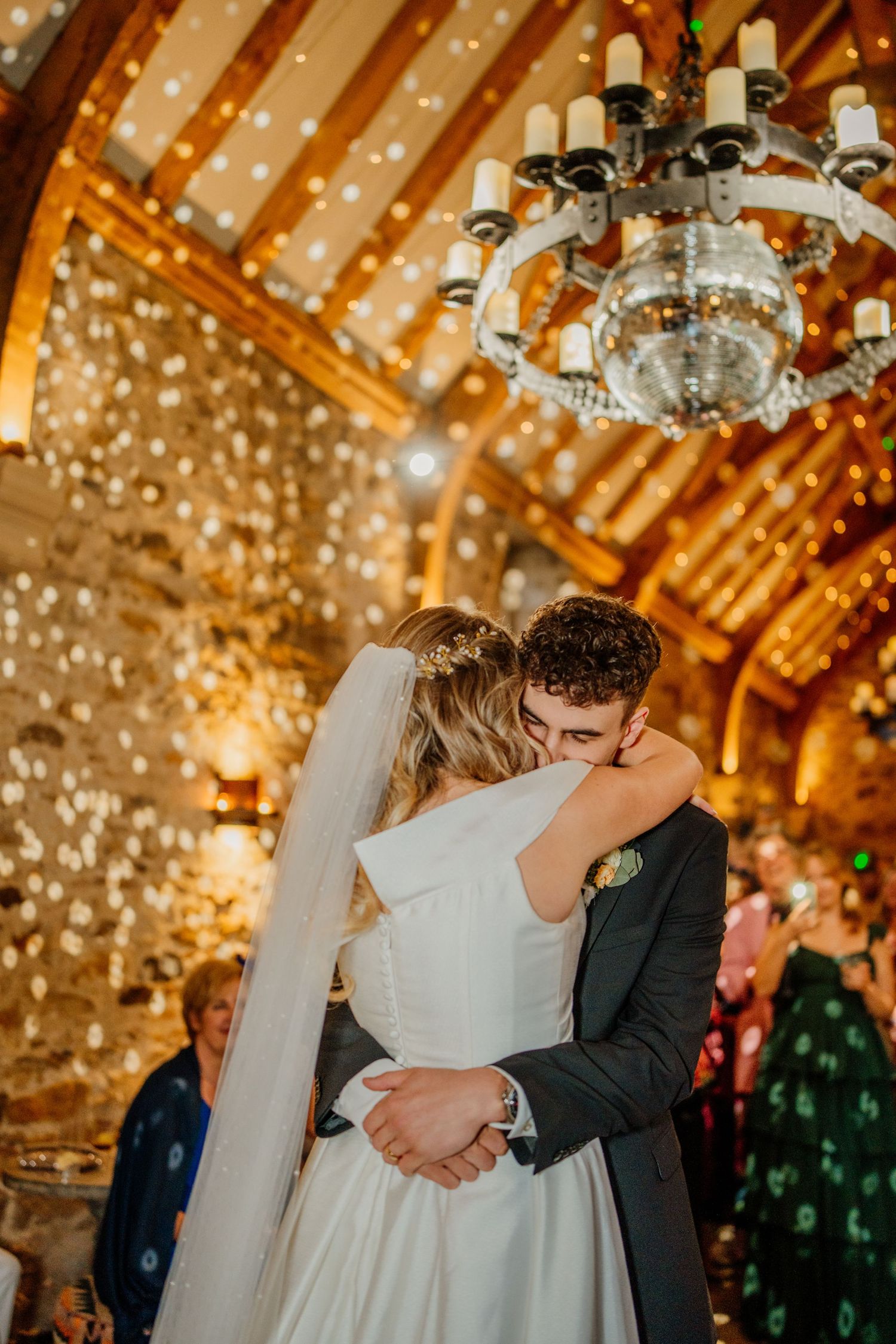 Couple shares romantic first dance under chandelier and fairy lights in rustic venue.