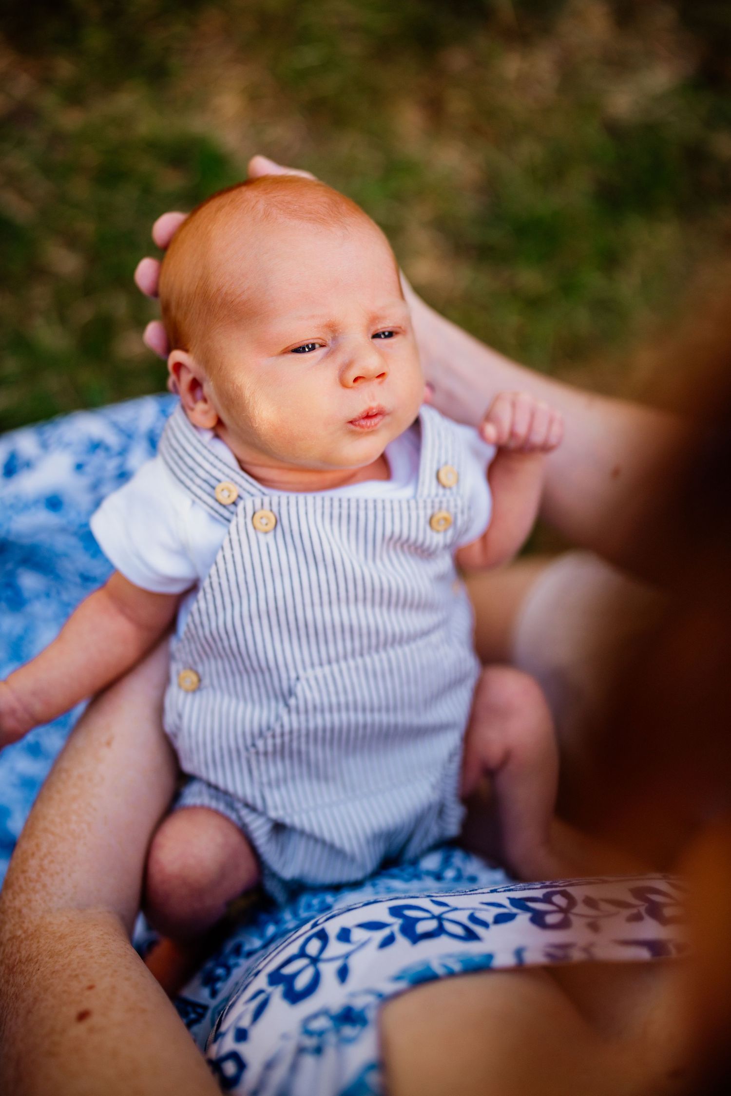 Newborn baby in striped romper being held outdoors on a blue floral blanket, looking alert and curious.