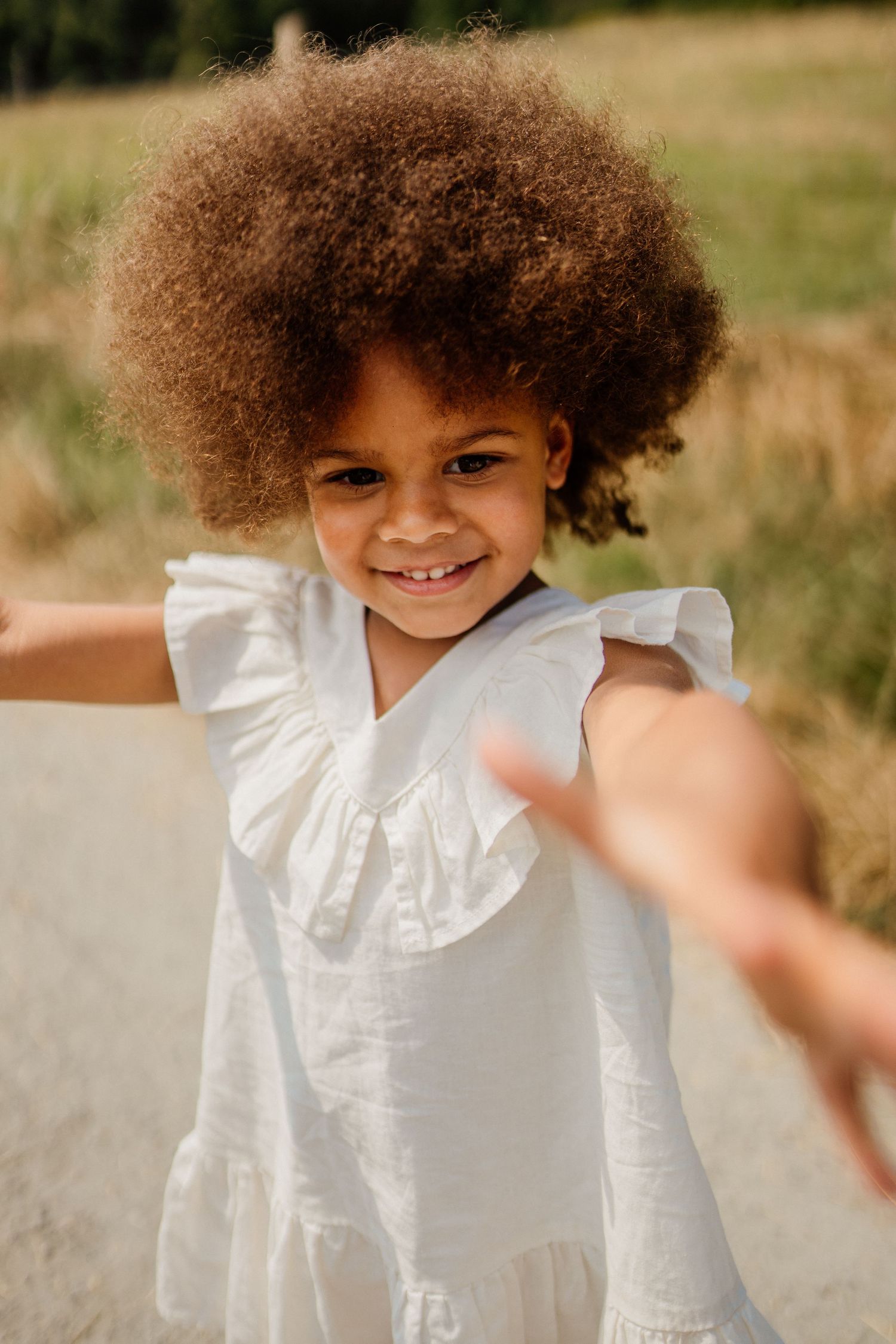 Child wearing white ruffled dress with natural curly hair playing outdoors in sunlit field.