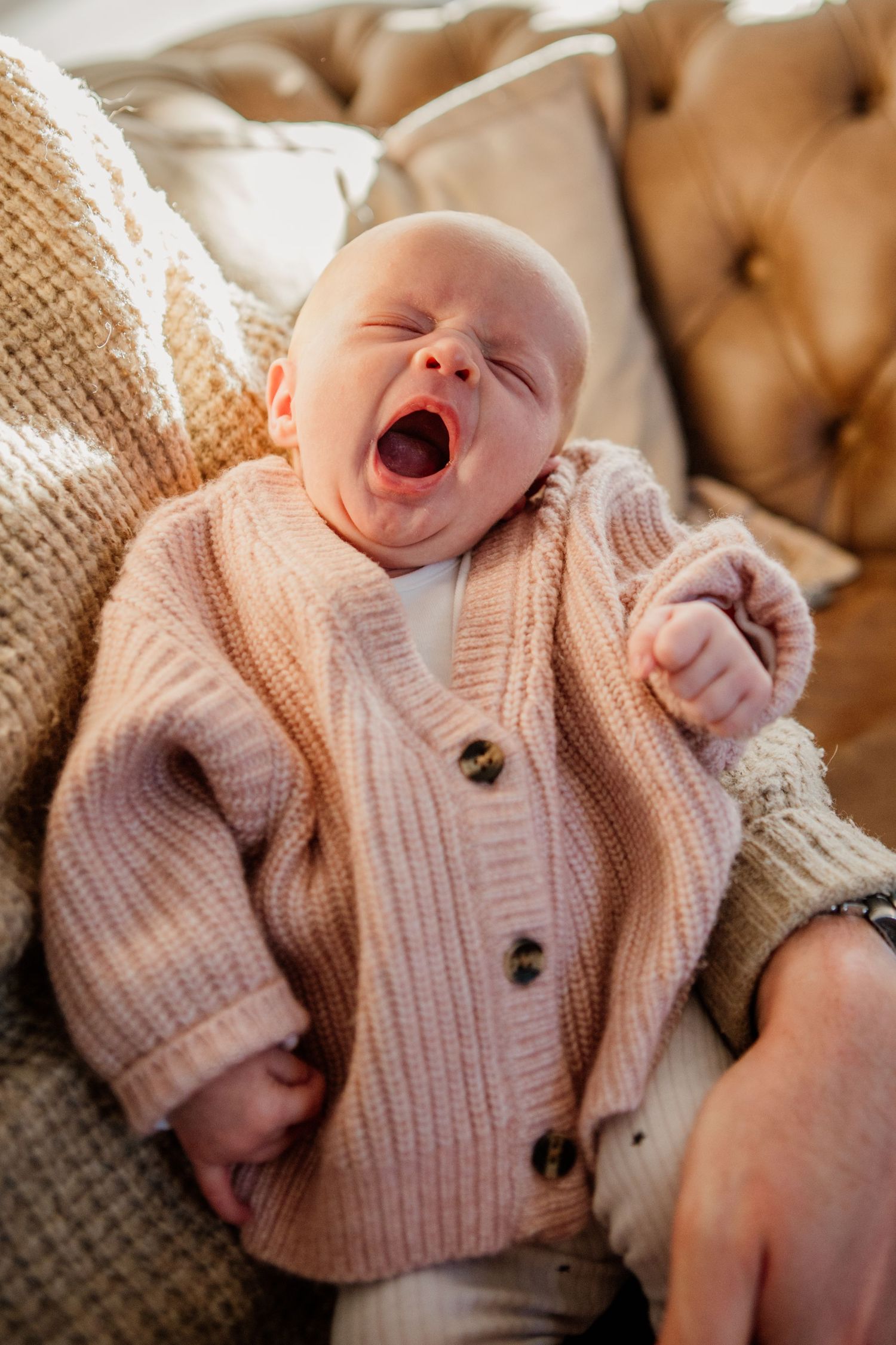 Baby wearing pink knit cardigan yawning while lying on textured fabric.