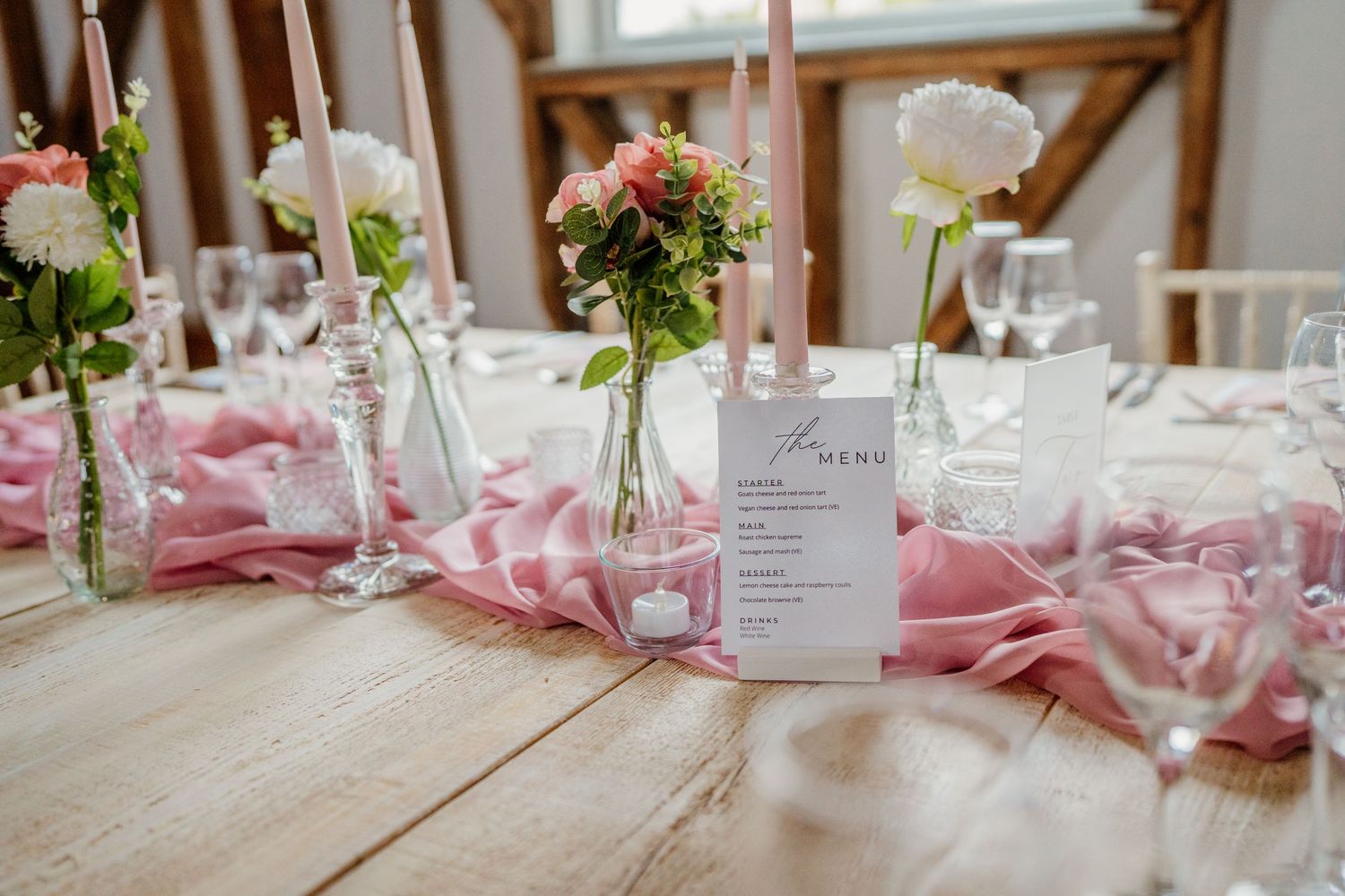 Elegant wedding reception table settings with pink accents, white linens, and floral centerpieces in a bright venue.