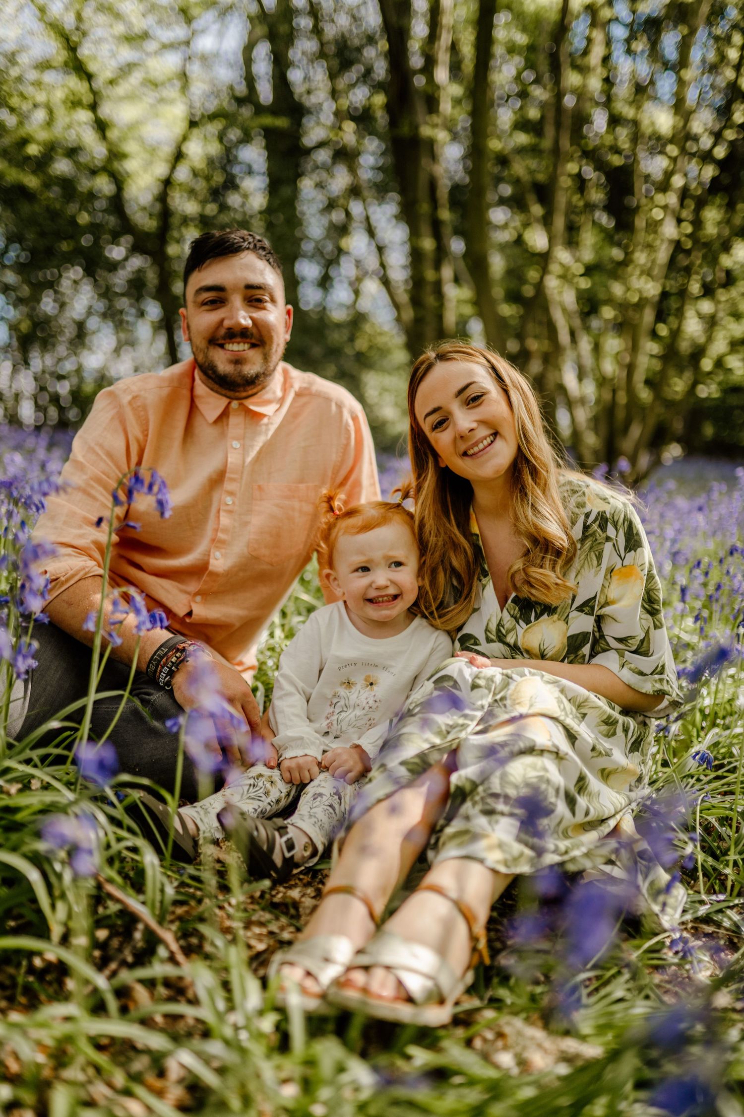 A family sits together in a blooming field of purple bluebells in Hertfordshire on a sunny spring day in the woods.