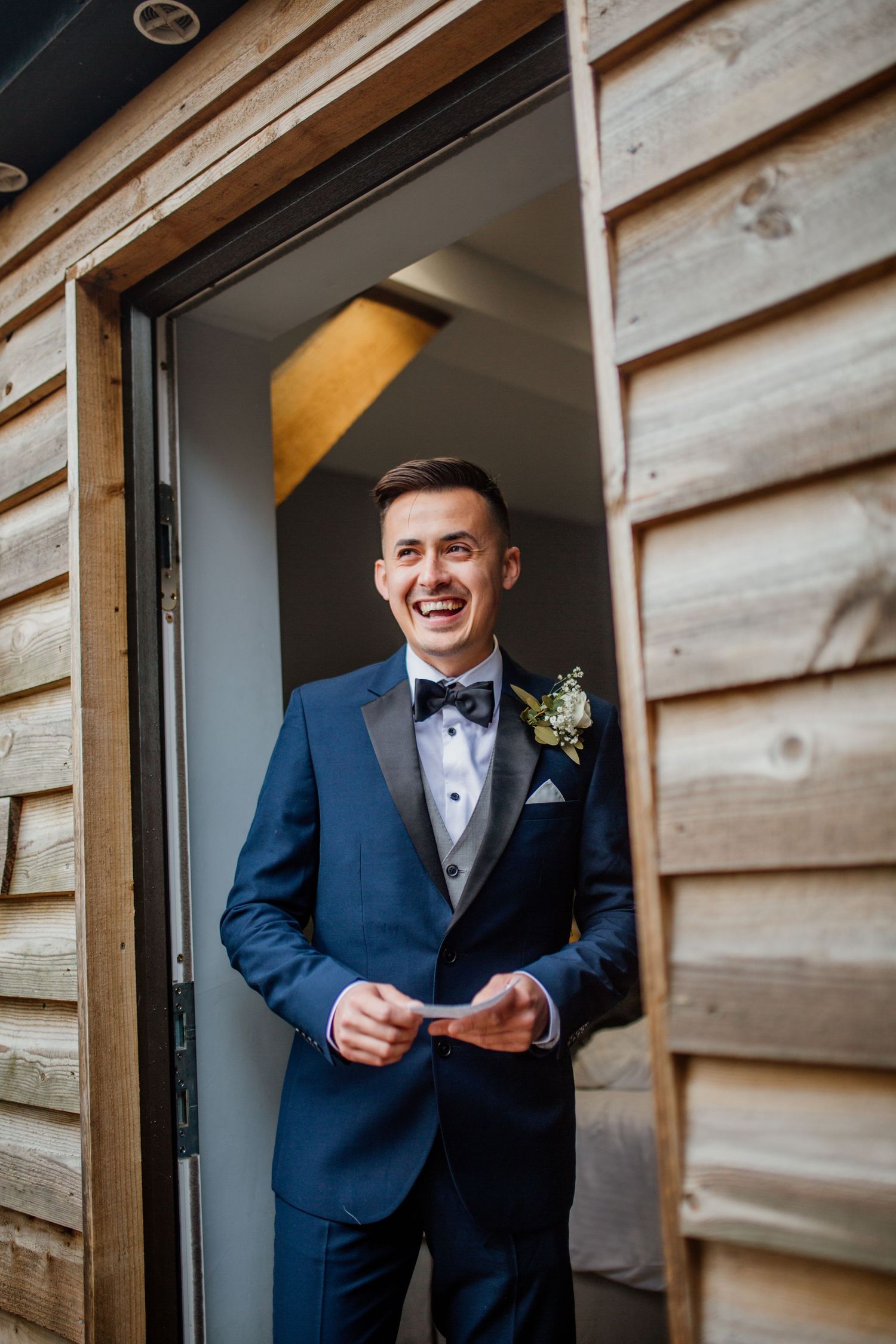 Groom in navy tuxedo reading letter in rustic wooden doorway.
