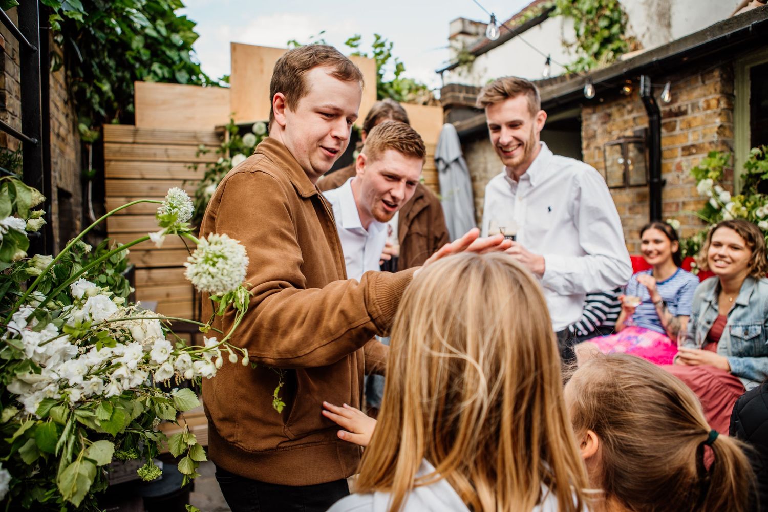 A group gathers in a garden patio setting with white flowers and greenery decorating the outdoor space.