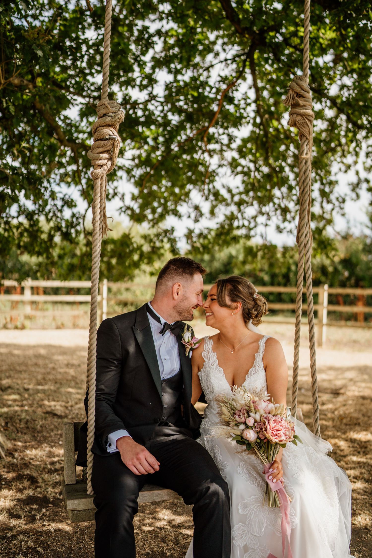 Newlyweds share tender moments while sitting on a rustic wooden swing under leafy trees.