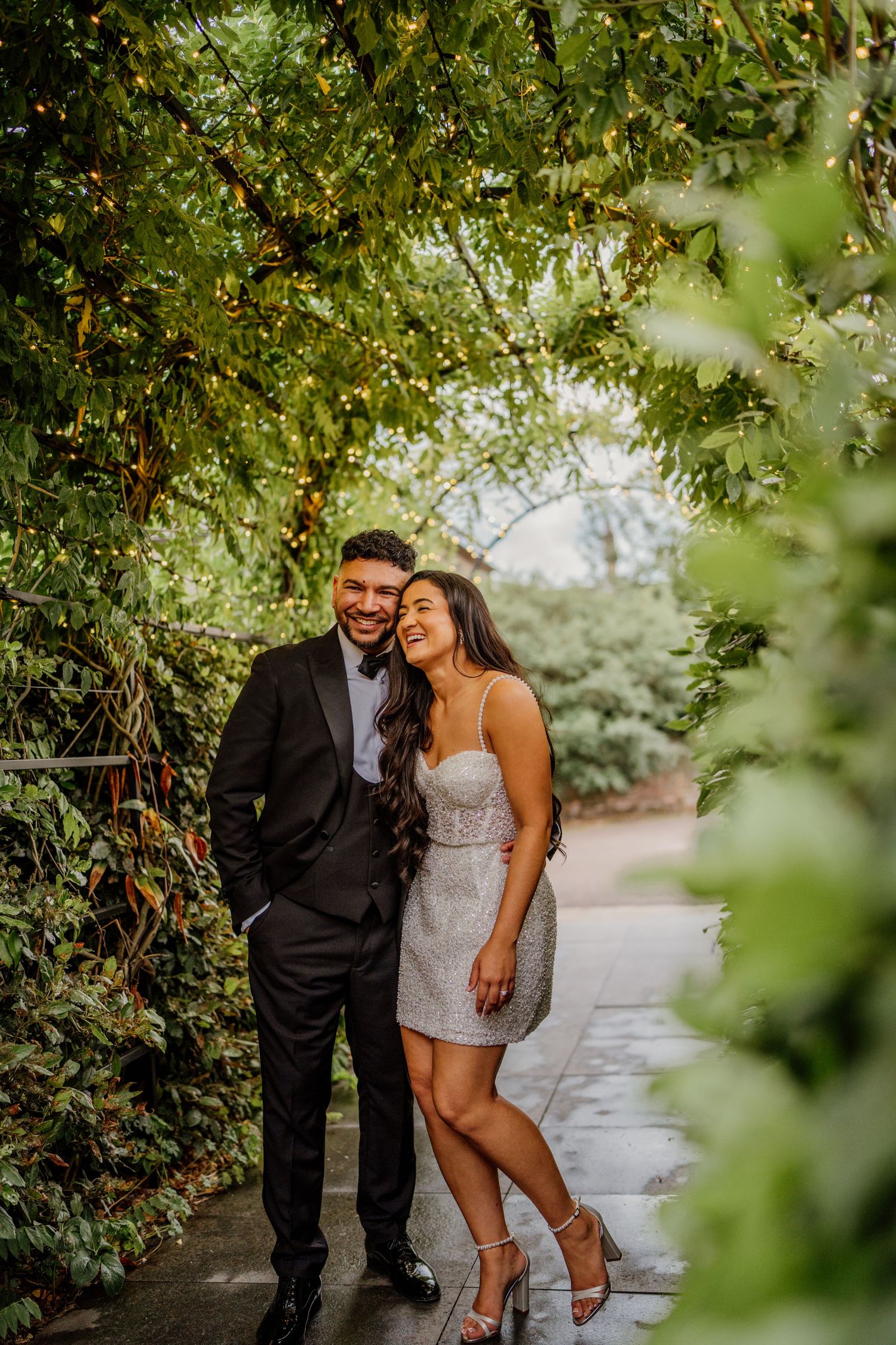 A couple shares a romantic moment on a garden path framed by lush greenery and natural light.