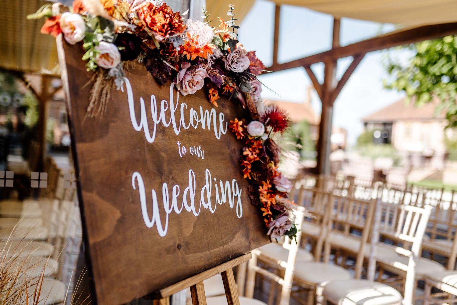 A rustic wooden welcome sign decorated with burgundy and blush floral arrangements at an outdoor wedding venue.