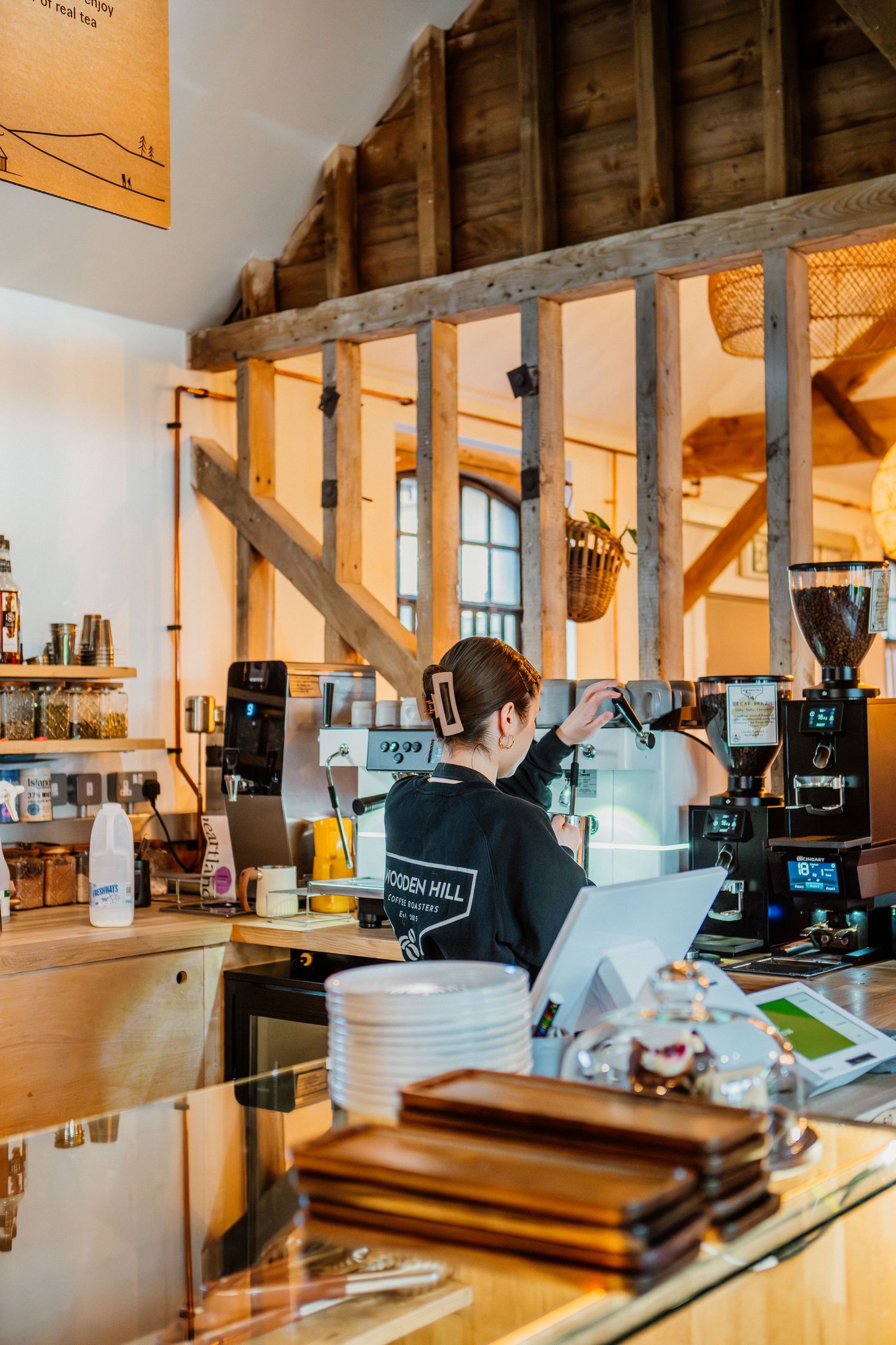 Barista working behind wooden counter at rustic coffee shop with exposed beams and warm lighting.