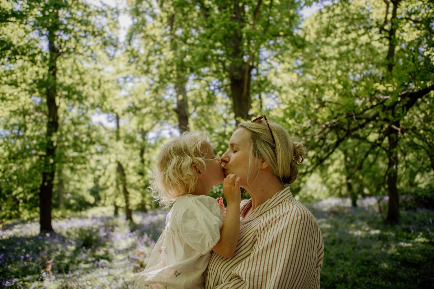 A tender outdoor moment shared in dappled sunlight beneath green trees.