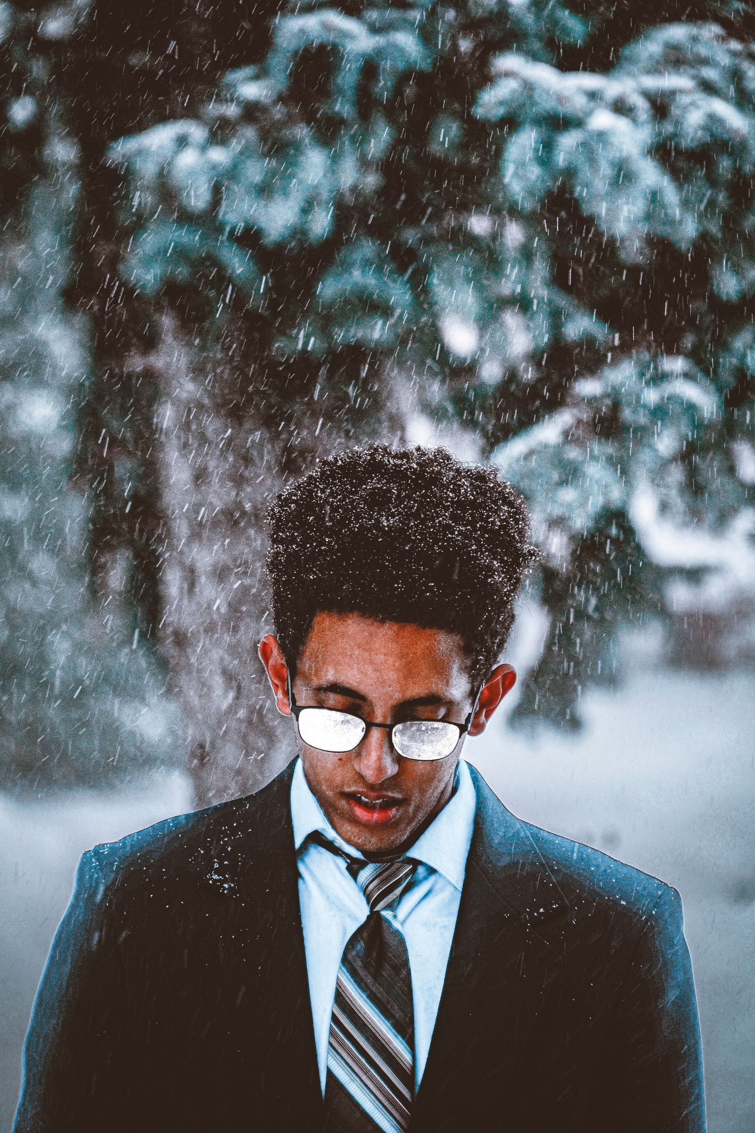 Professional wearing glasses and striped tie looks down while standing outdoors with snowy winter trees in background.