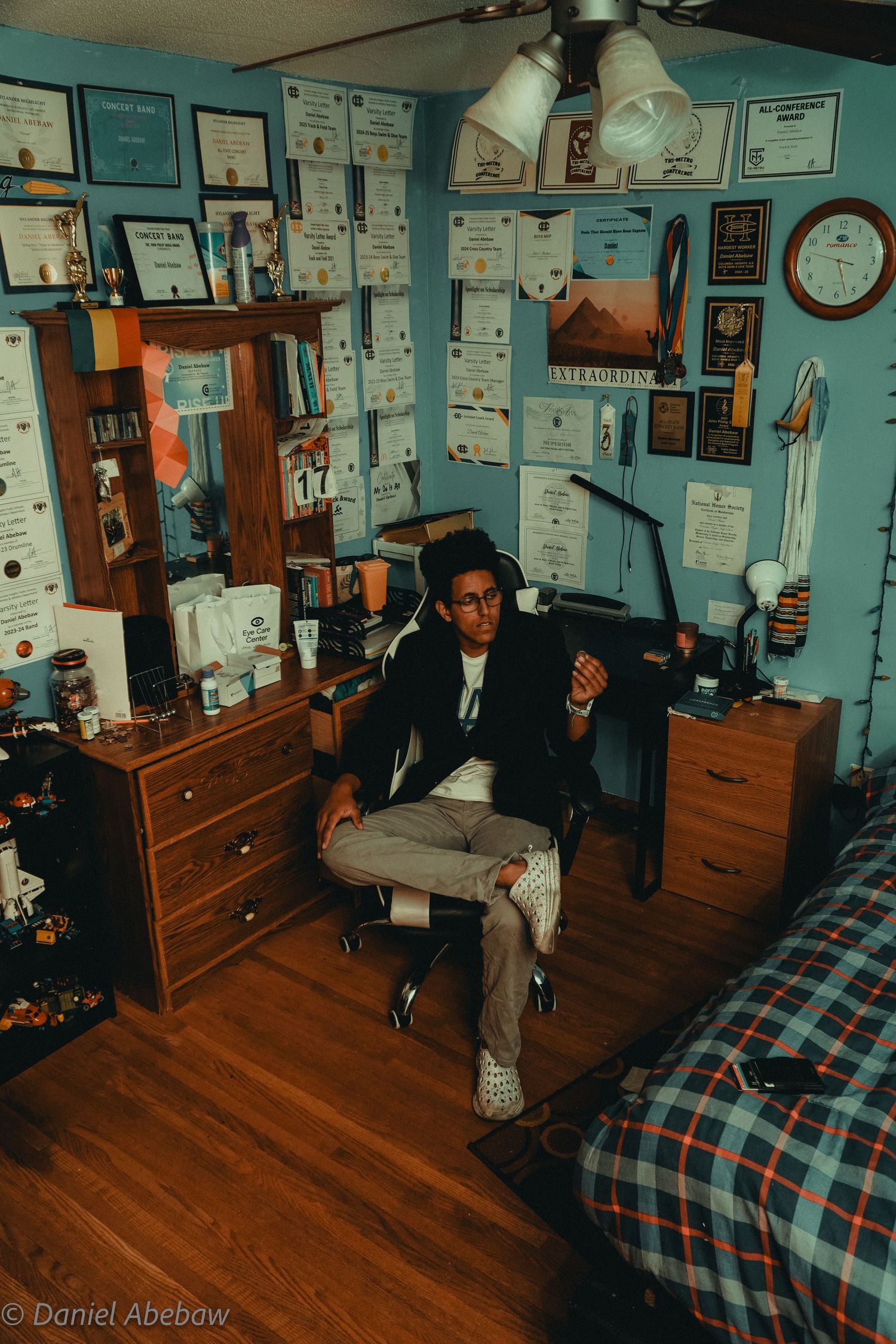 Young man sitting in office chair surrounded by awards and certificates on blue walls in a decorated bedroom.