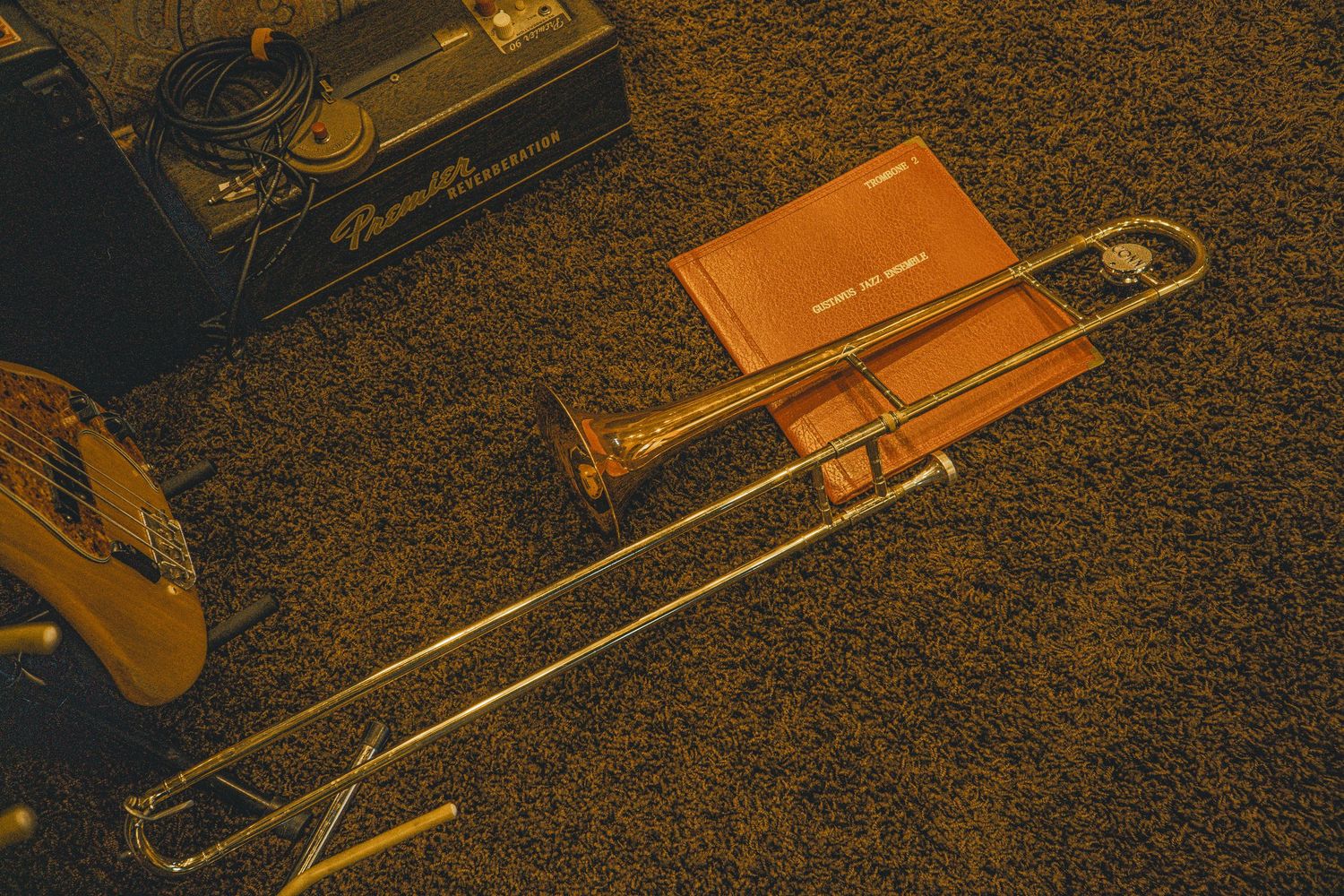 A vintage brass trombone resting on carpet beside sheet music and an amplifier, bathed in warm golden light.