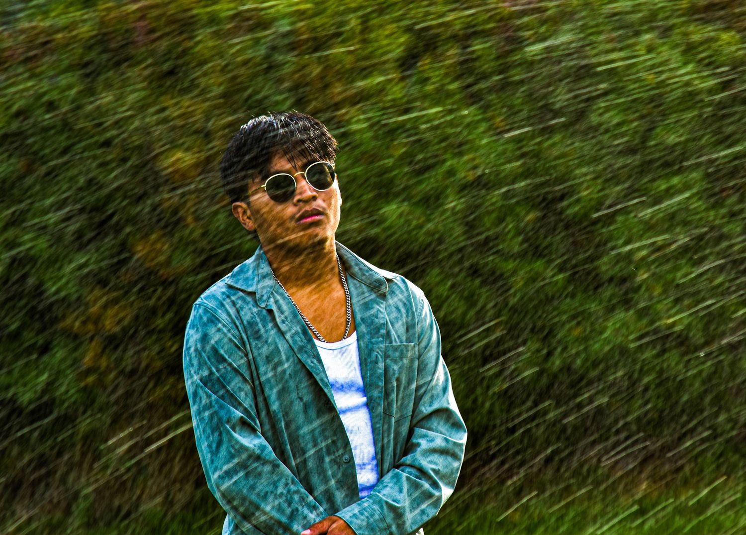 Figure in sunglasses and a blue jacket against lush green foliage behind sprinklers.