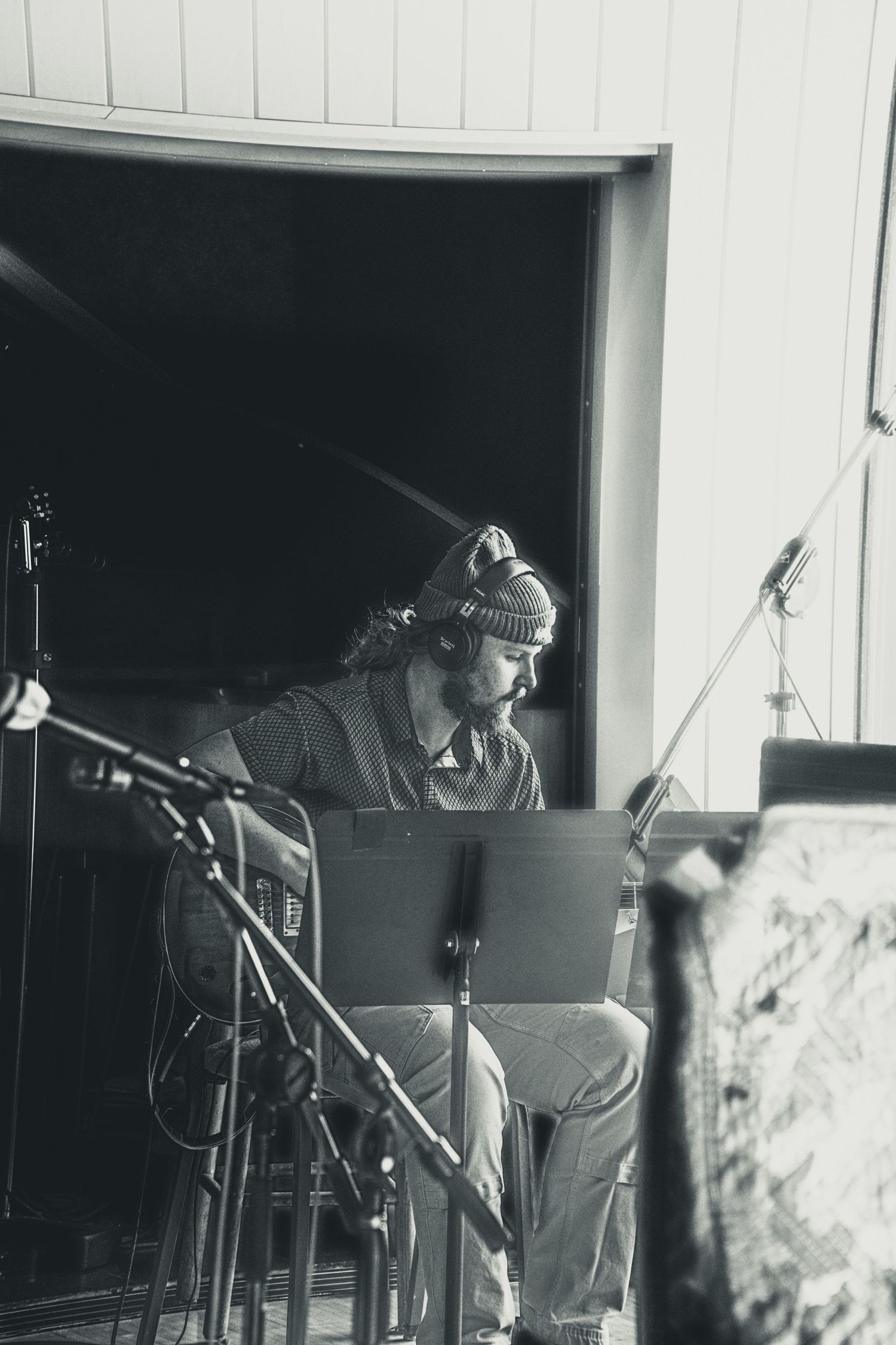 Black and white photo of a musician wearing a beanie, seated with sheet music stand and microphones in a recording studio.