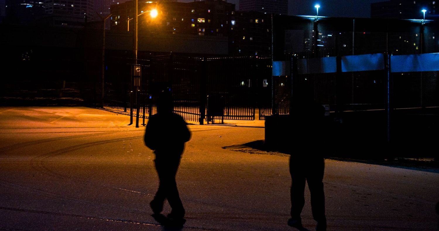 People walking in silhouette on a dimly lit street at night with street lamps glowing.