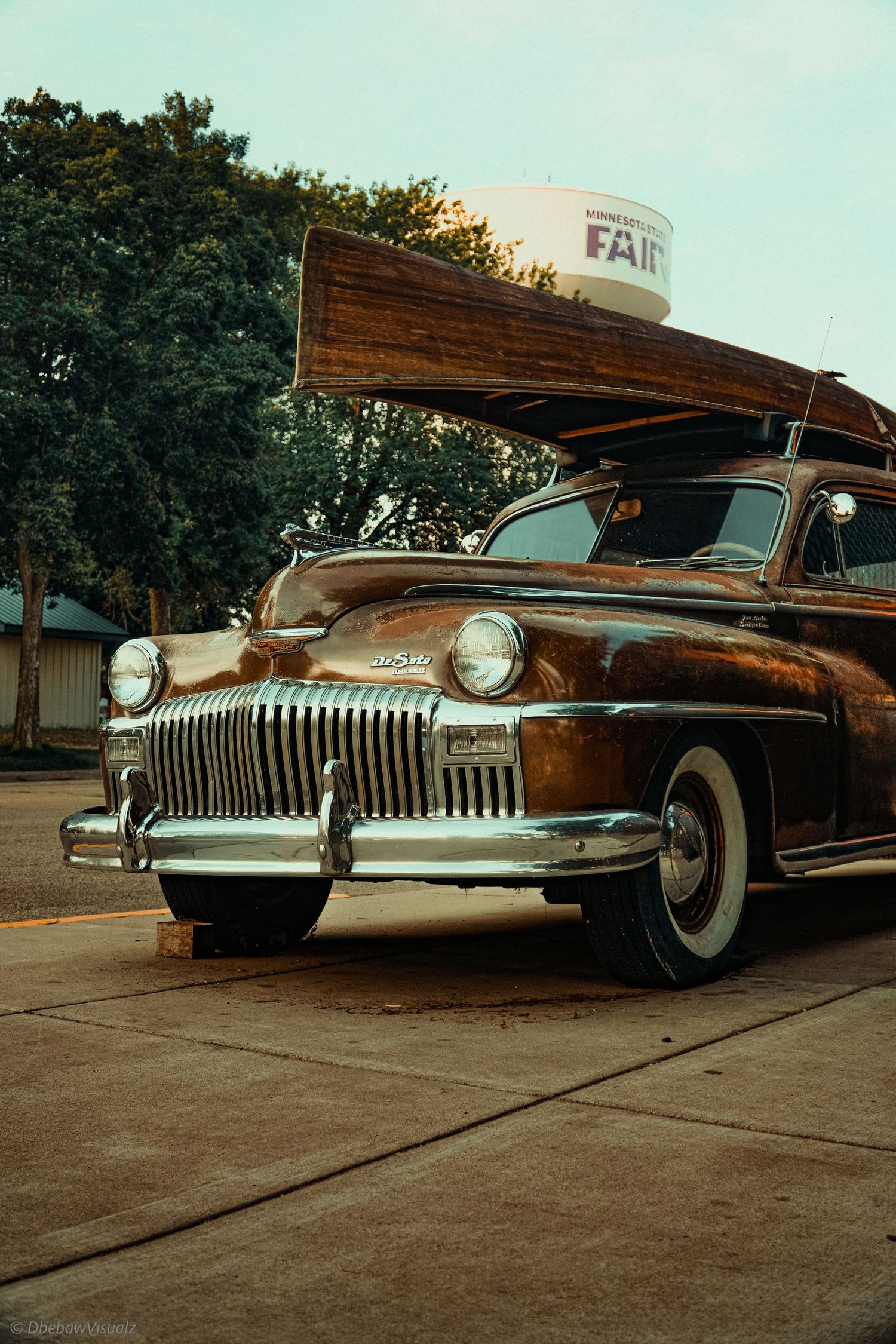 Vintage DeSoto classic car with wood-topped roof rack and whitewall tires parked near a fairground water tower.