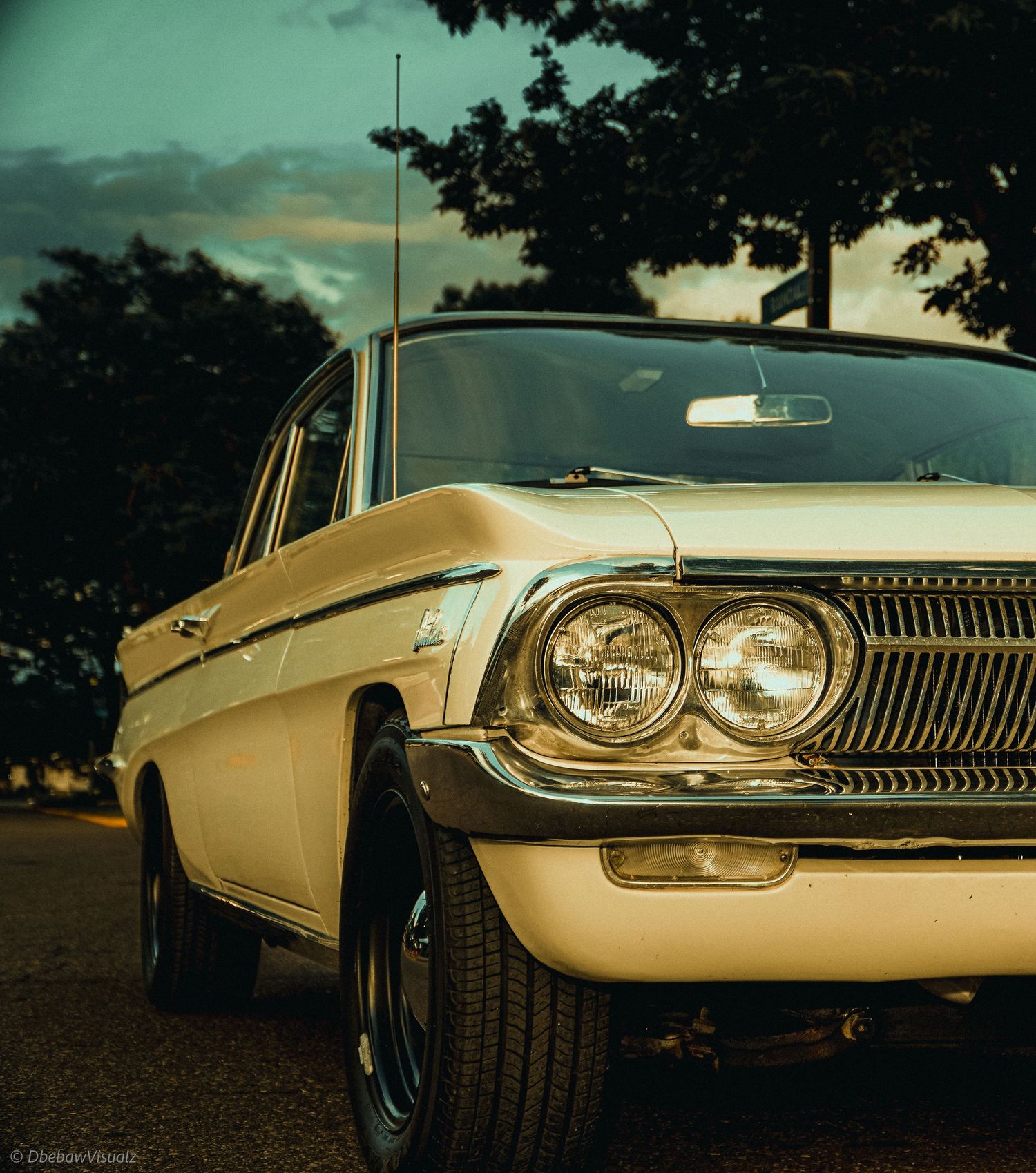 Vintage cream-colored classic car parked outdoors at dusk with dramatic sky and trees in background.