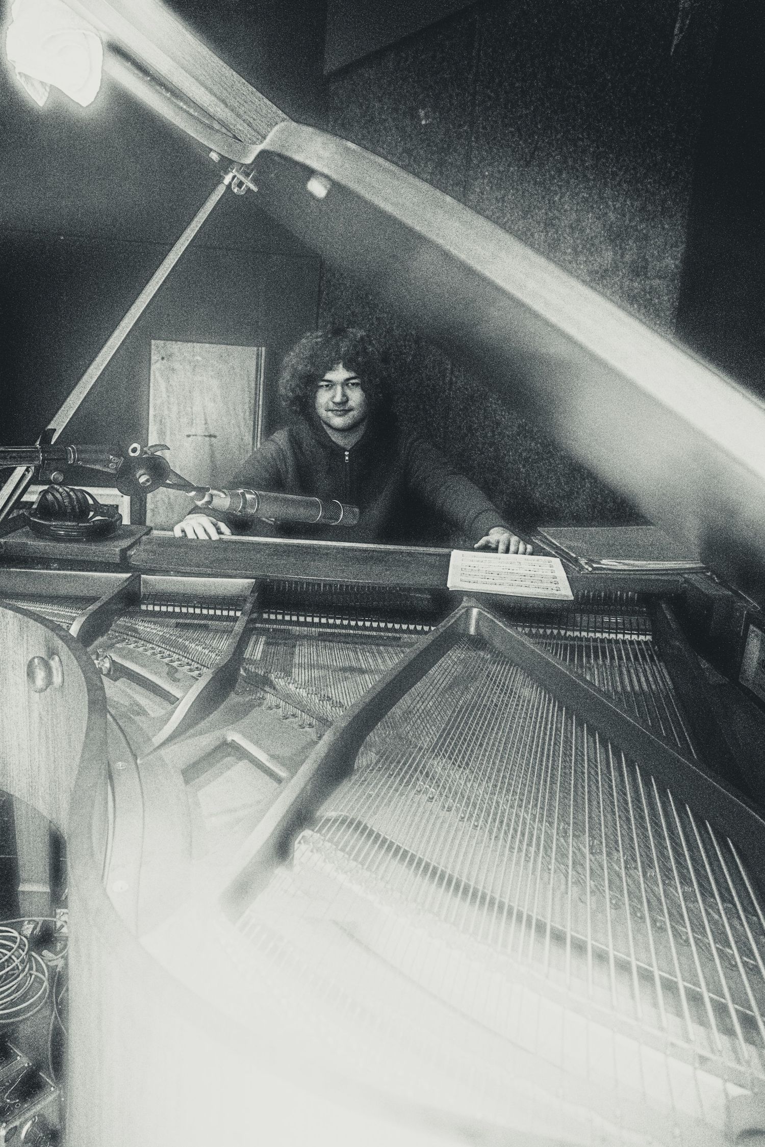 Black and white photo of a curly-haired musician smiling behind an open grand piano in a studio.