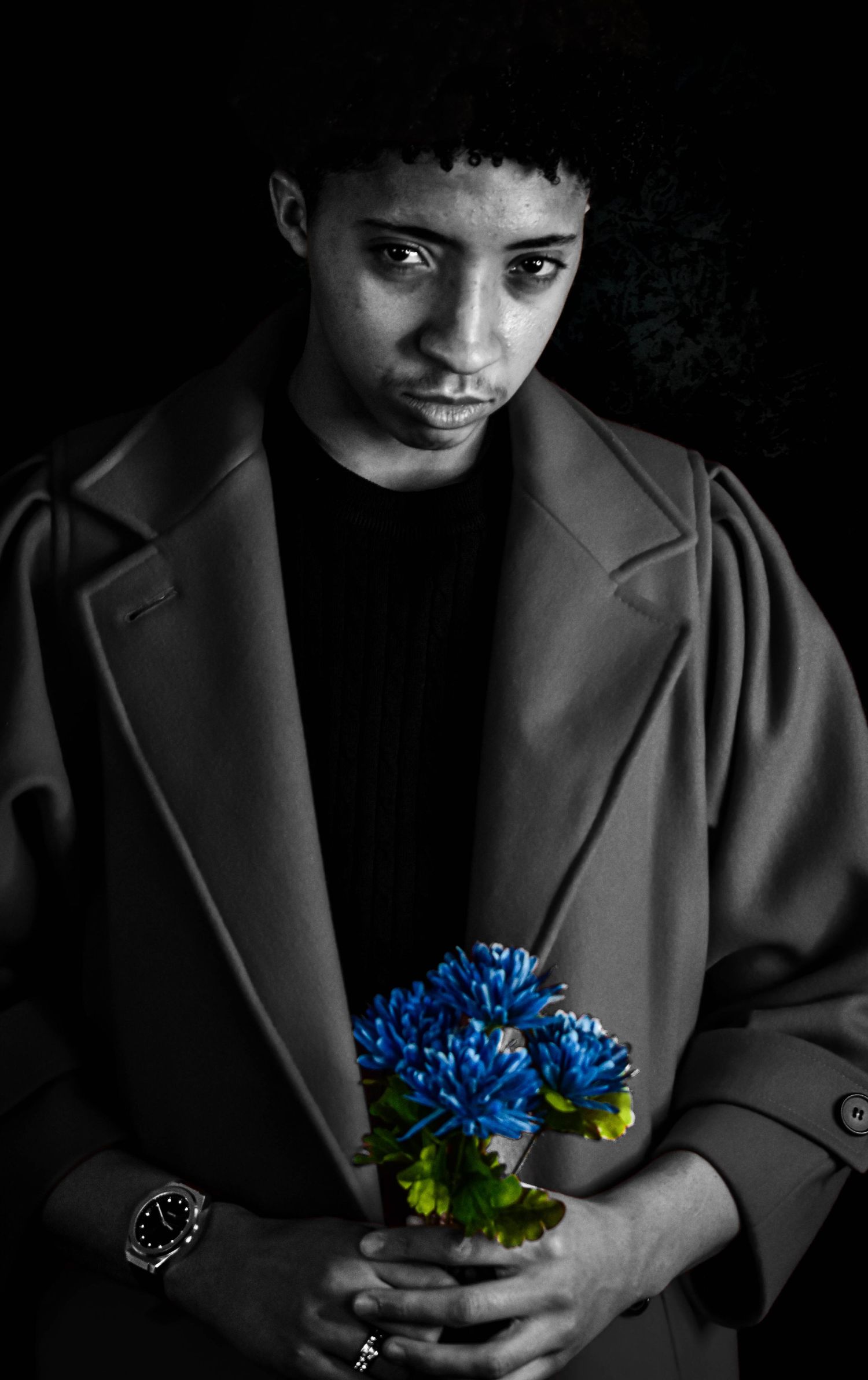 Dramatic portrait holding bright blue flowers against dark background in formal coat.