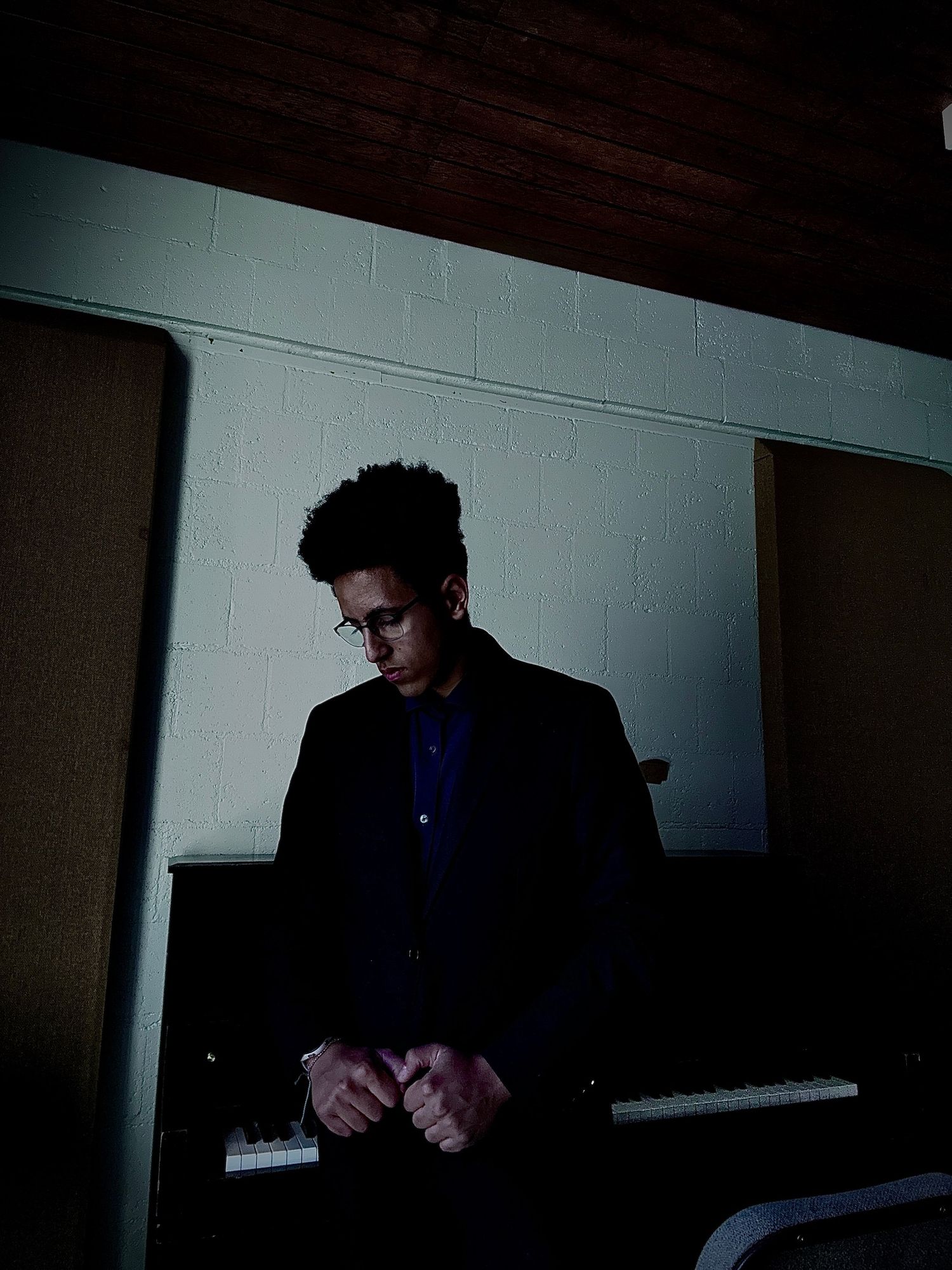 A man in a dark blazer stands pensively near a keyboard in a dimly lit studio room.