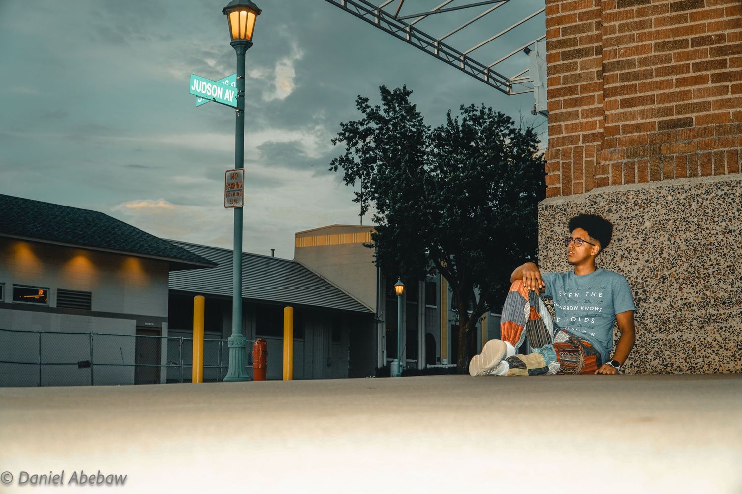 Young man sitting against a brick wall at dusk near a street lamp in an urban setting, wearing a graphic tee.