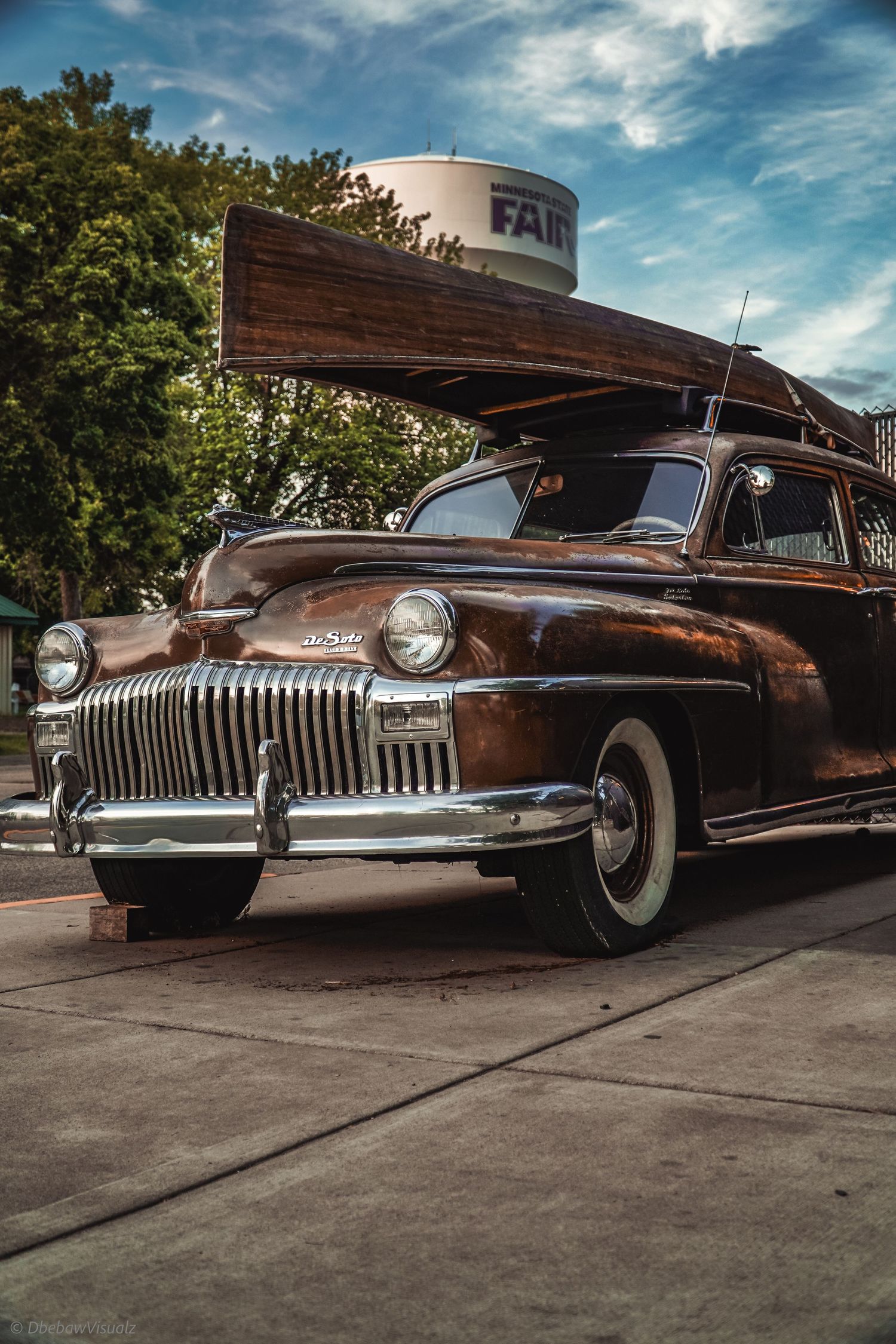 Vintage DeSoto classic car in brown with chrome grille and whitewall tires, carrying a wooden canoe on roof rack outdoors.