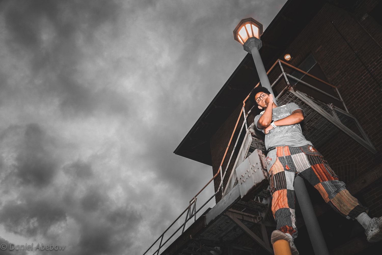 Person standing tall near a lamp post on a structure under dramatic cloudy skies in selective color photography.