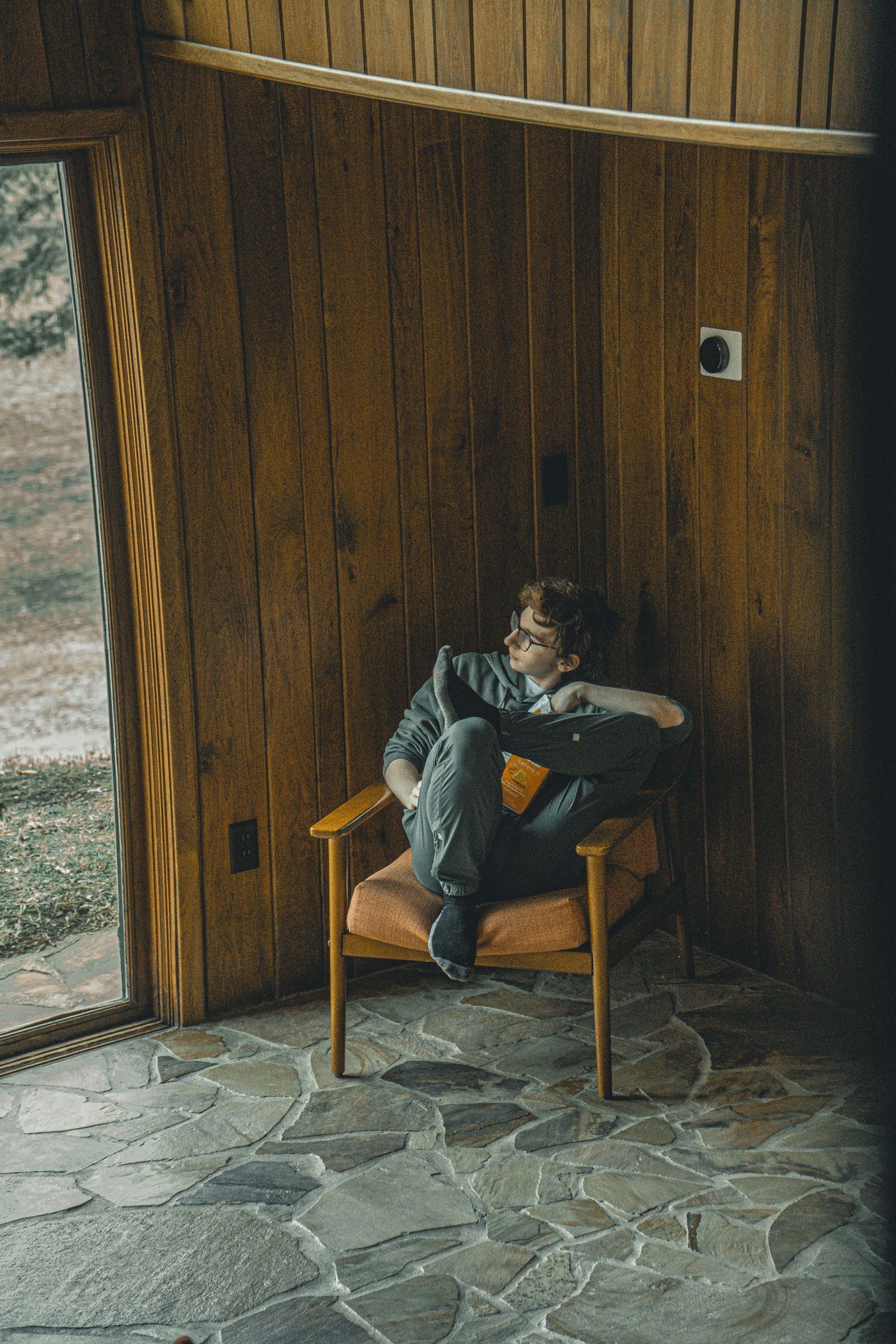 Young man with glasses relaxing in a mid-century modern leather chair in a wood-paneled room with stone tile floor.