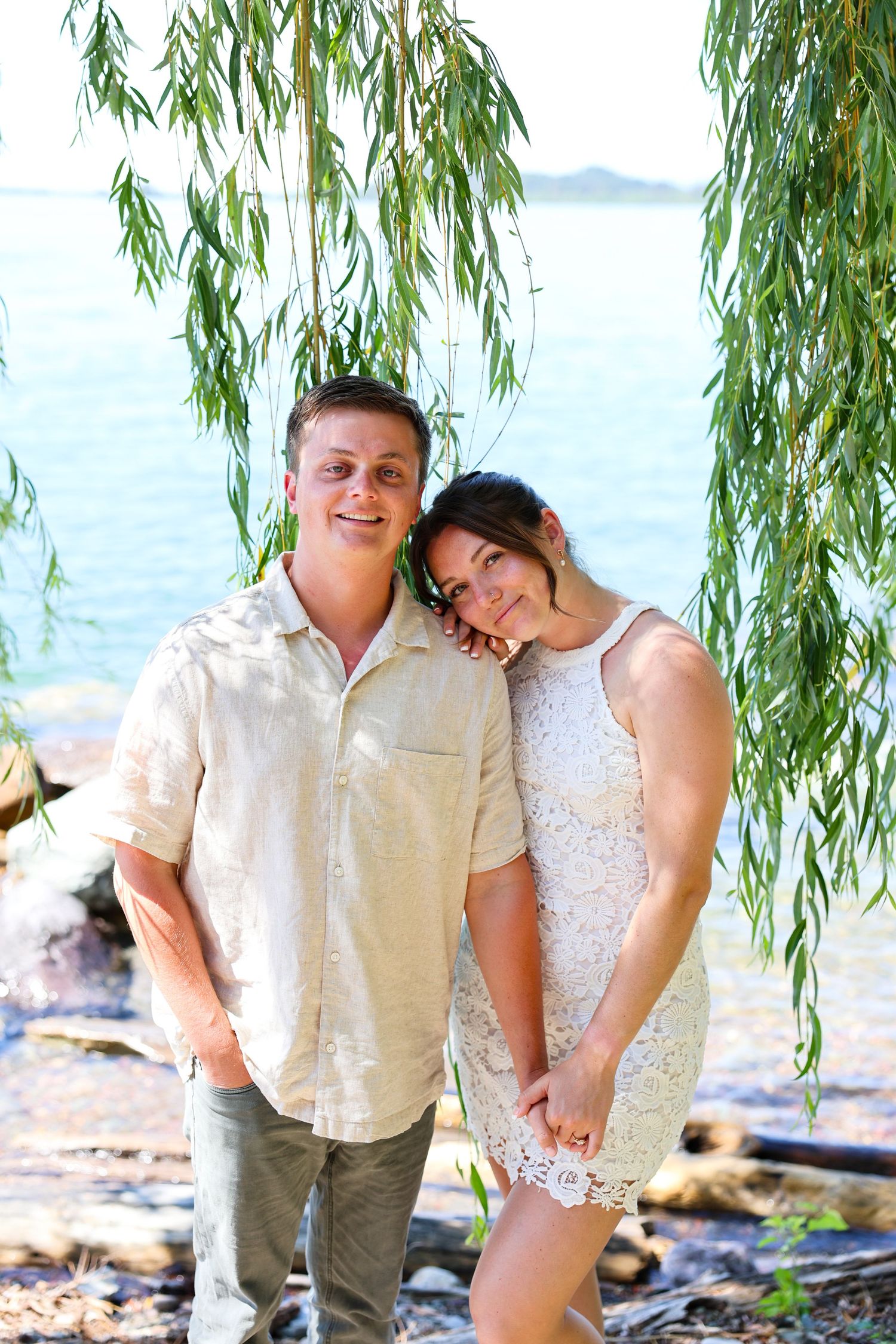 Engaged couple pose in front of the lake with beautiful greenery surrounding them.