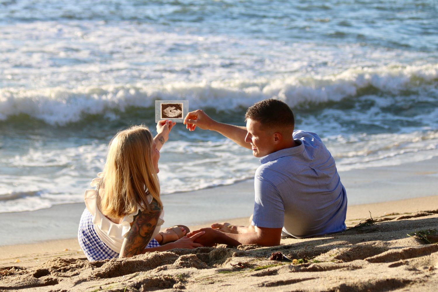 Couple holds up a sonogram in front of the ocean, announcing they are expecting a baby girl on Valentines Day.
