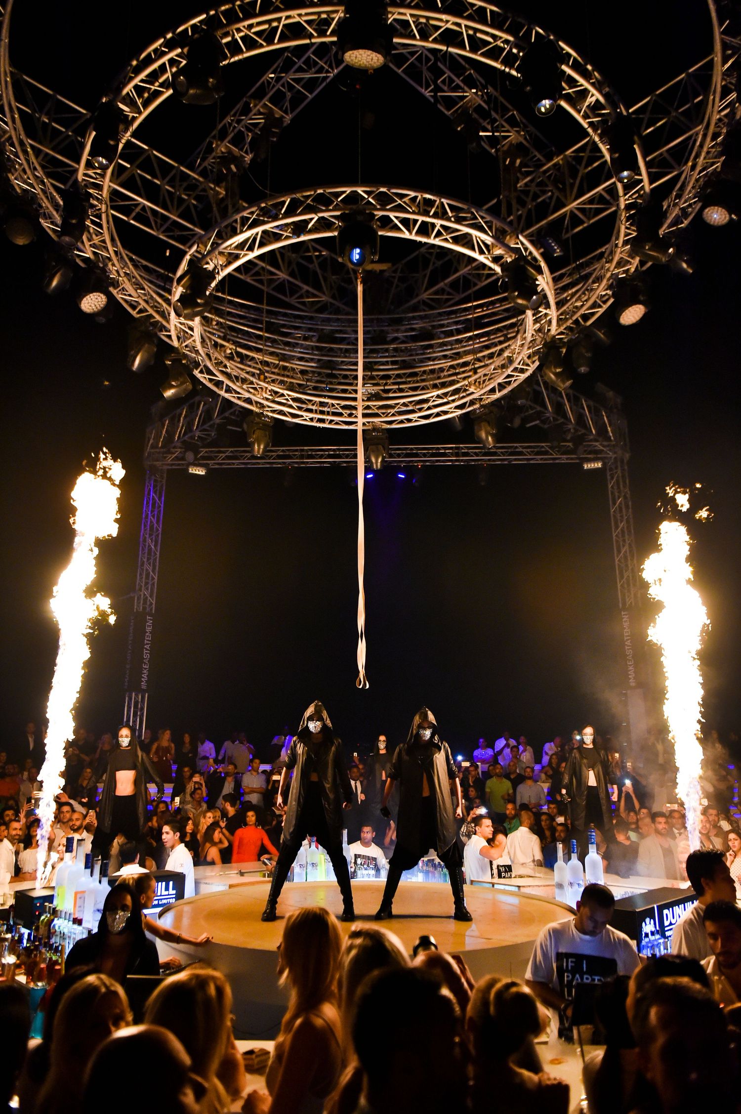 Indoor event venue with circular truss rigging and pyrotechnic effects shooting flames upward.
