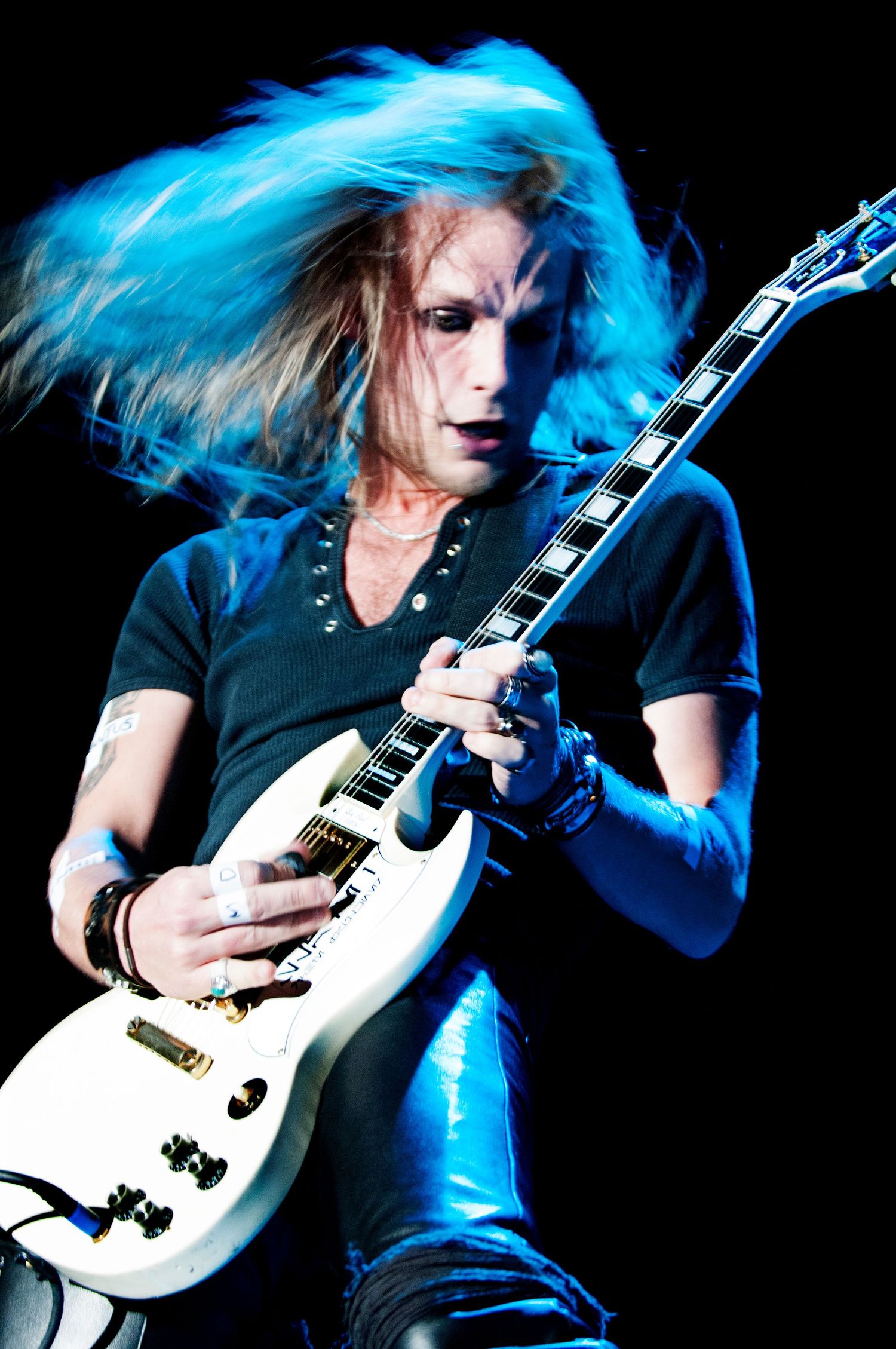 A guitarist with bright blue hair performs energetically on stage with a white electric guitar under dramatic lighting. Event photographer