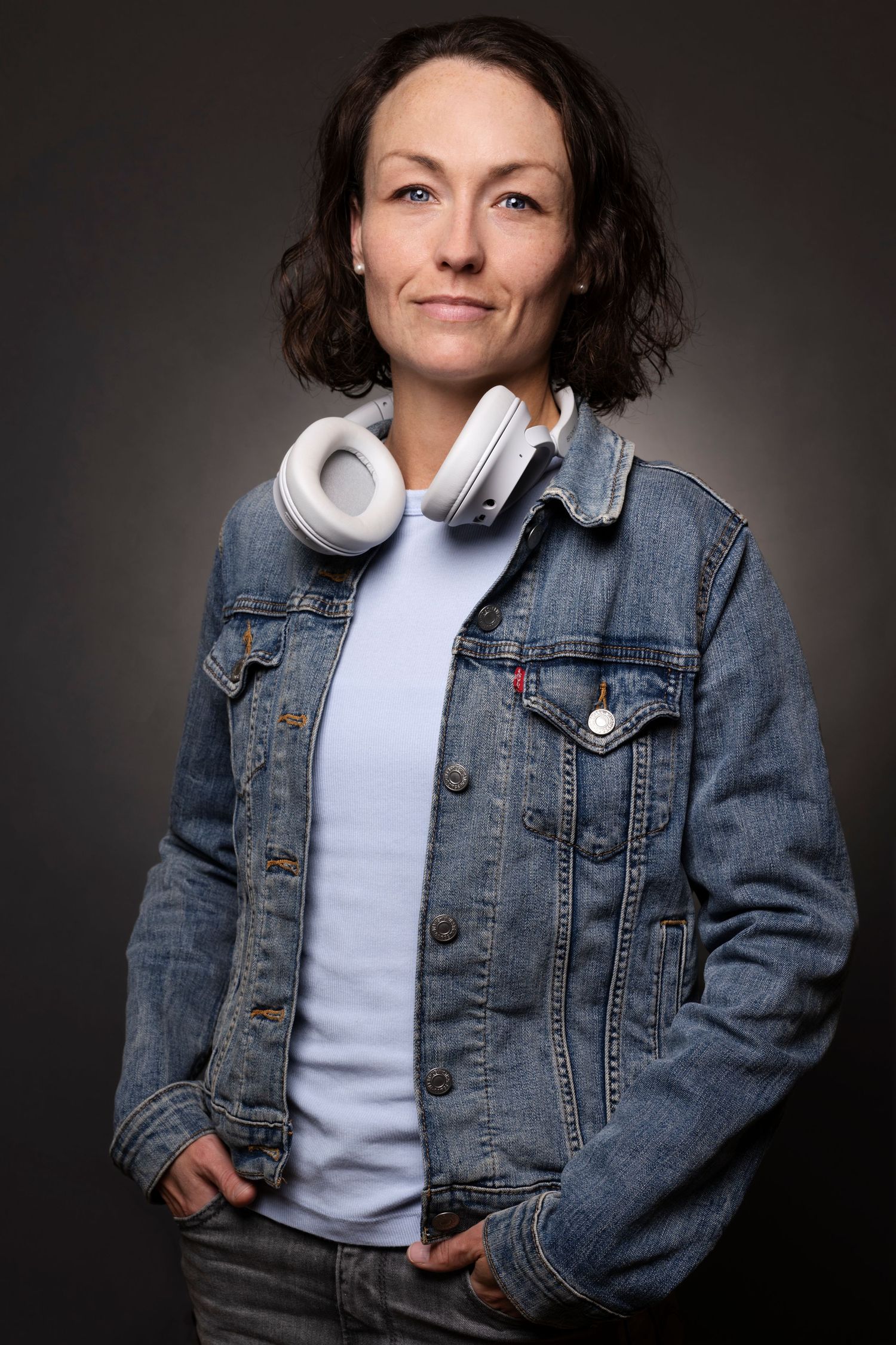 Person in denim jacket and white headphones poses against dark background. Portrait Photographer in Ontario