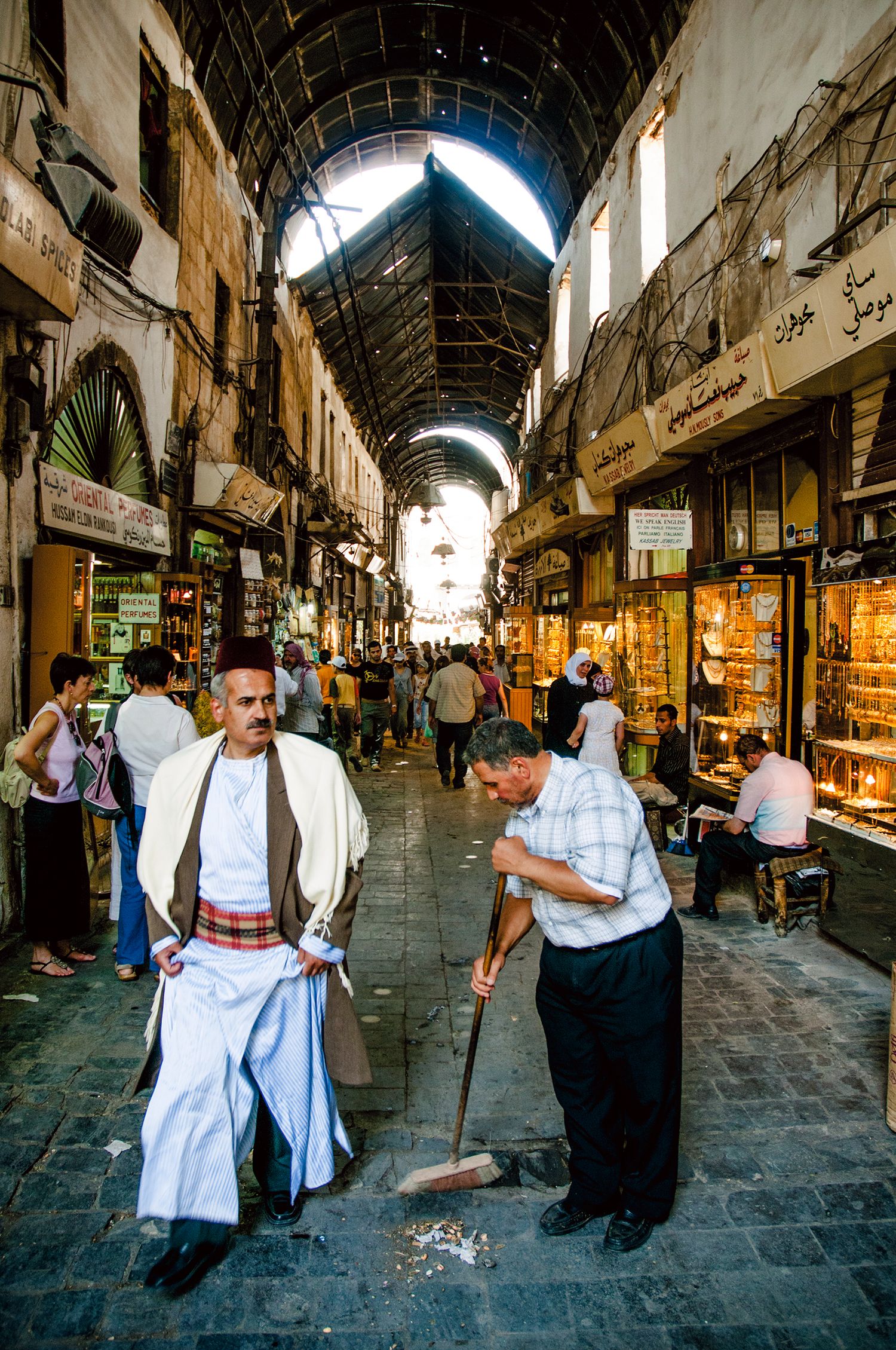 People walk through a historic covered market with stone arches and shops along both sides in Damascus, Syria.  Souk Al Hamidiya