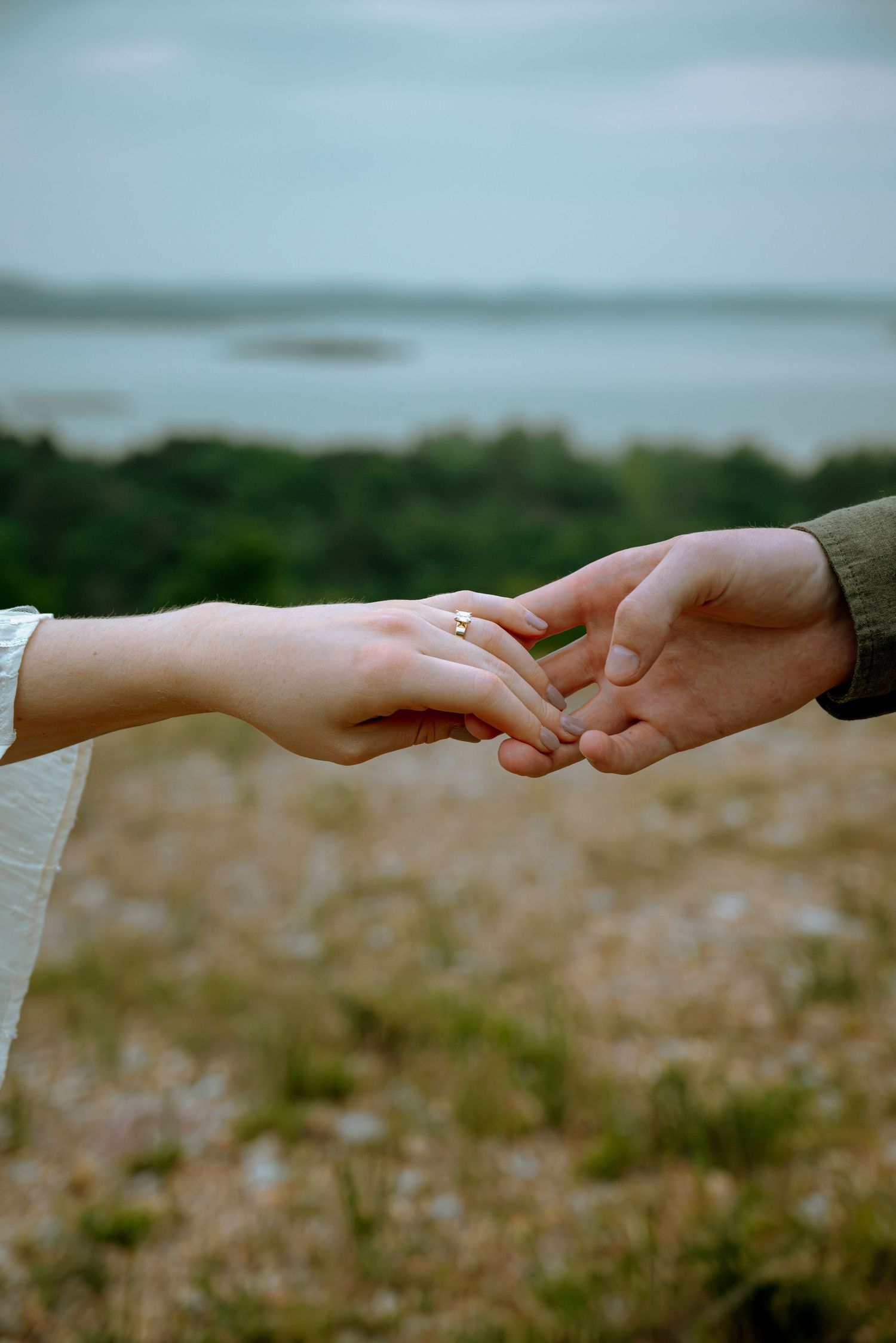 Close-up of newly engaged couple holding hands displaying diamond engagement ring with lake in background.