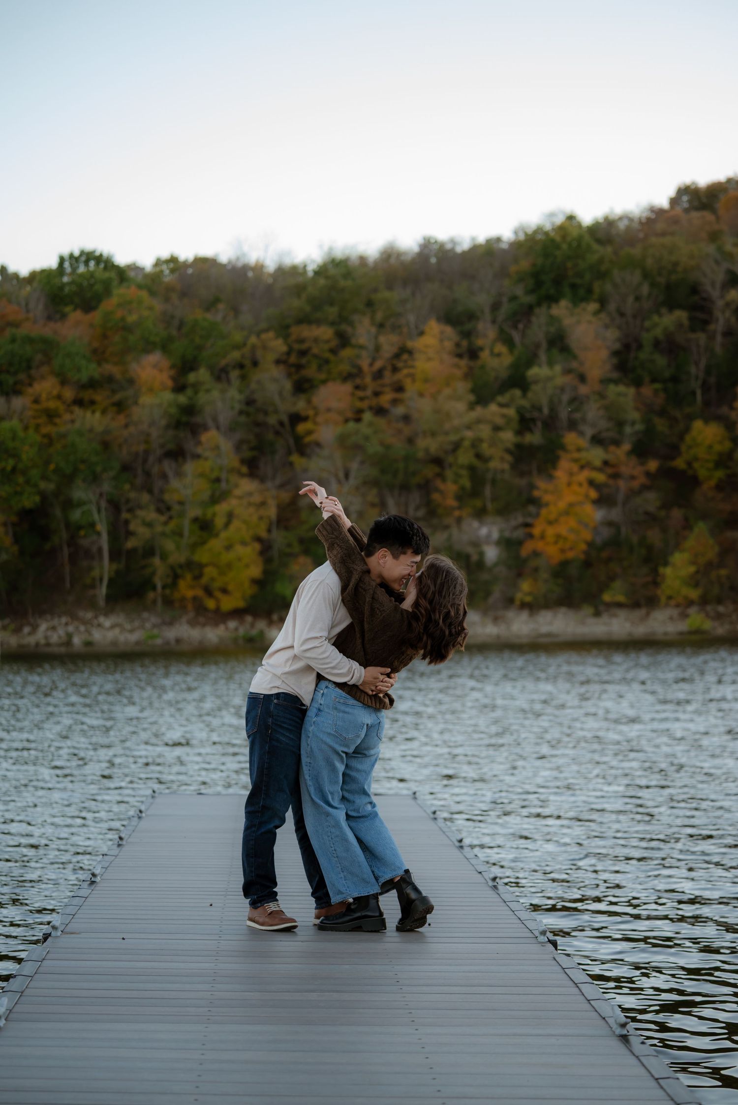 Couple sharing a romantic kiss on a lake dock in autumn, surrounded by colorful fall foliage and calm water.