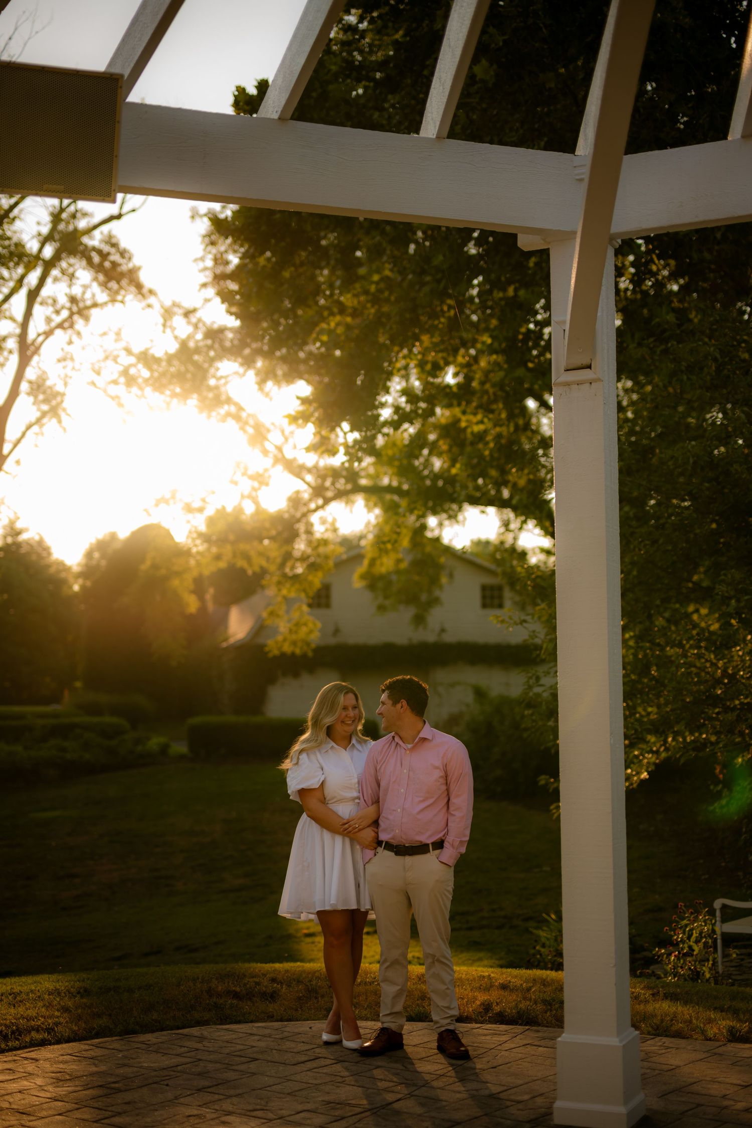 Couple sharing a romantic moment under a white pergola at golden hour, surrounded by lush green trees.