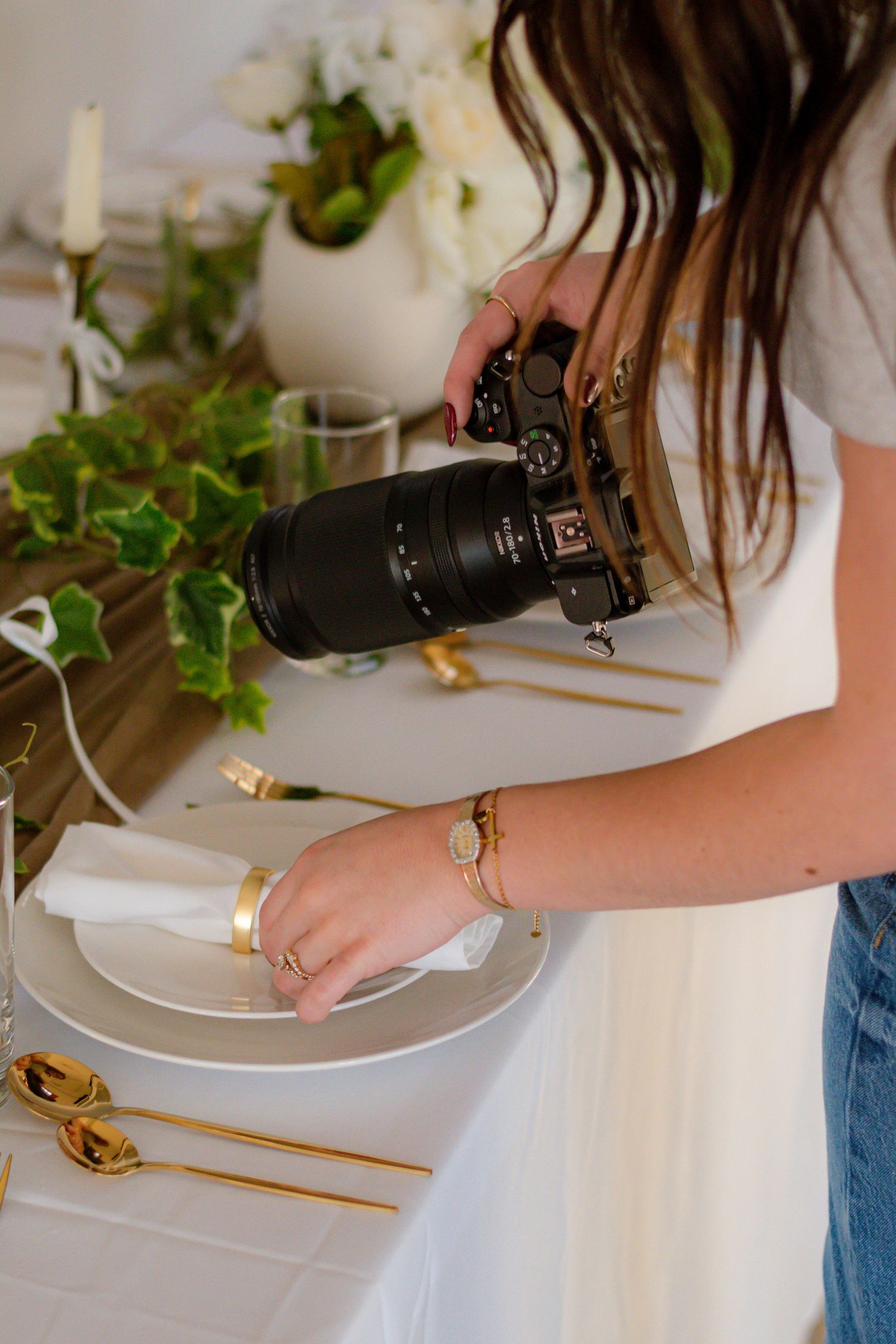 Female photographer leaning over elegant white table setting with gold accents, capturing detail shots with DSLR.