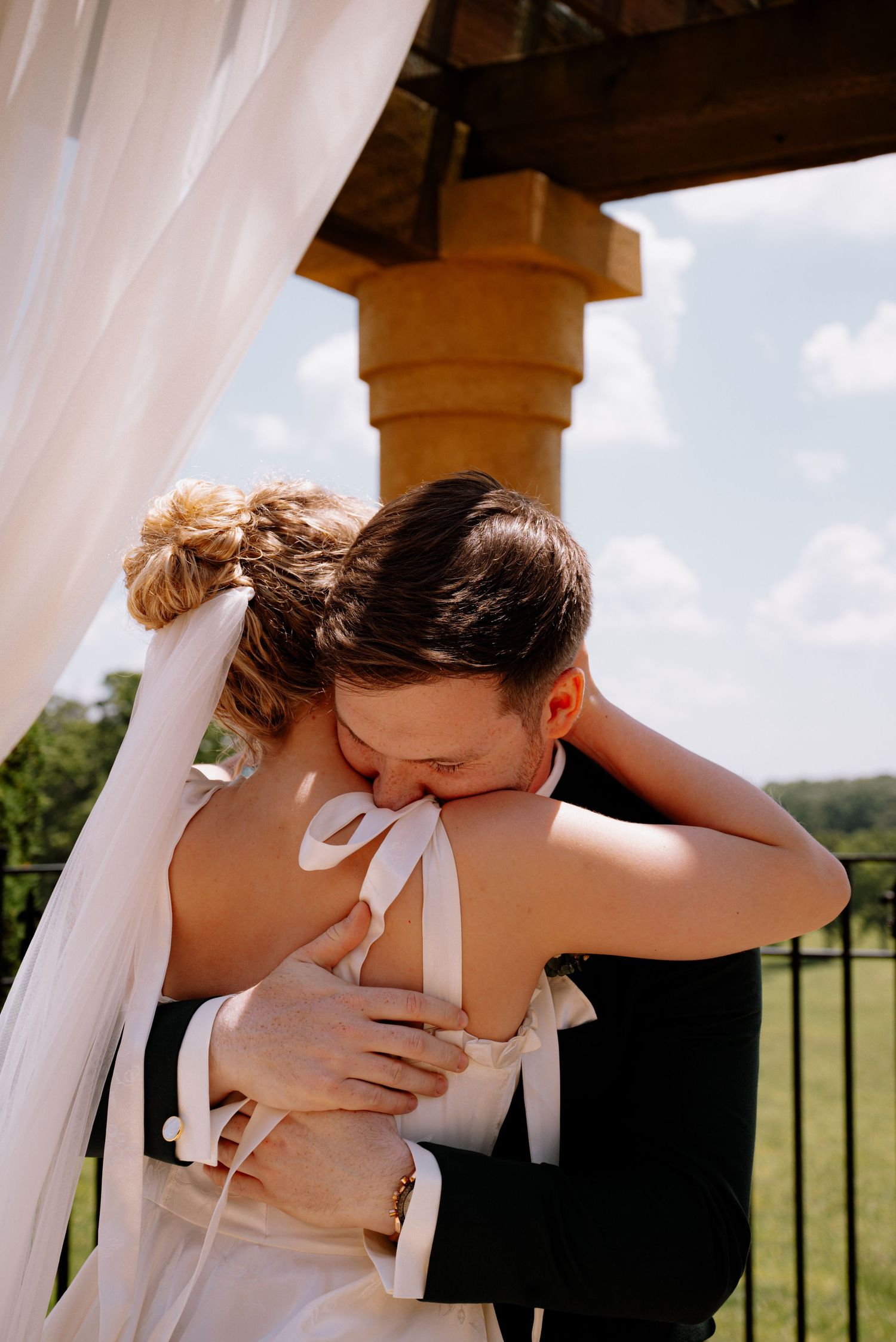 Bride and groom share an emotional first embrace at outdoor wedding altar under white draped canopy.